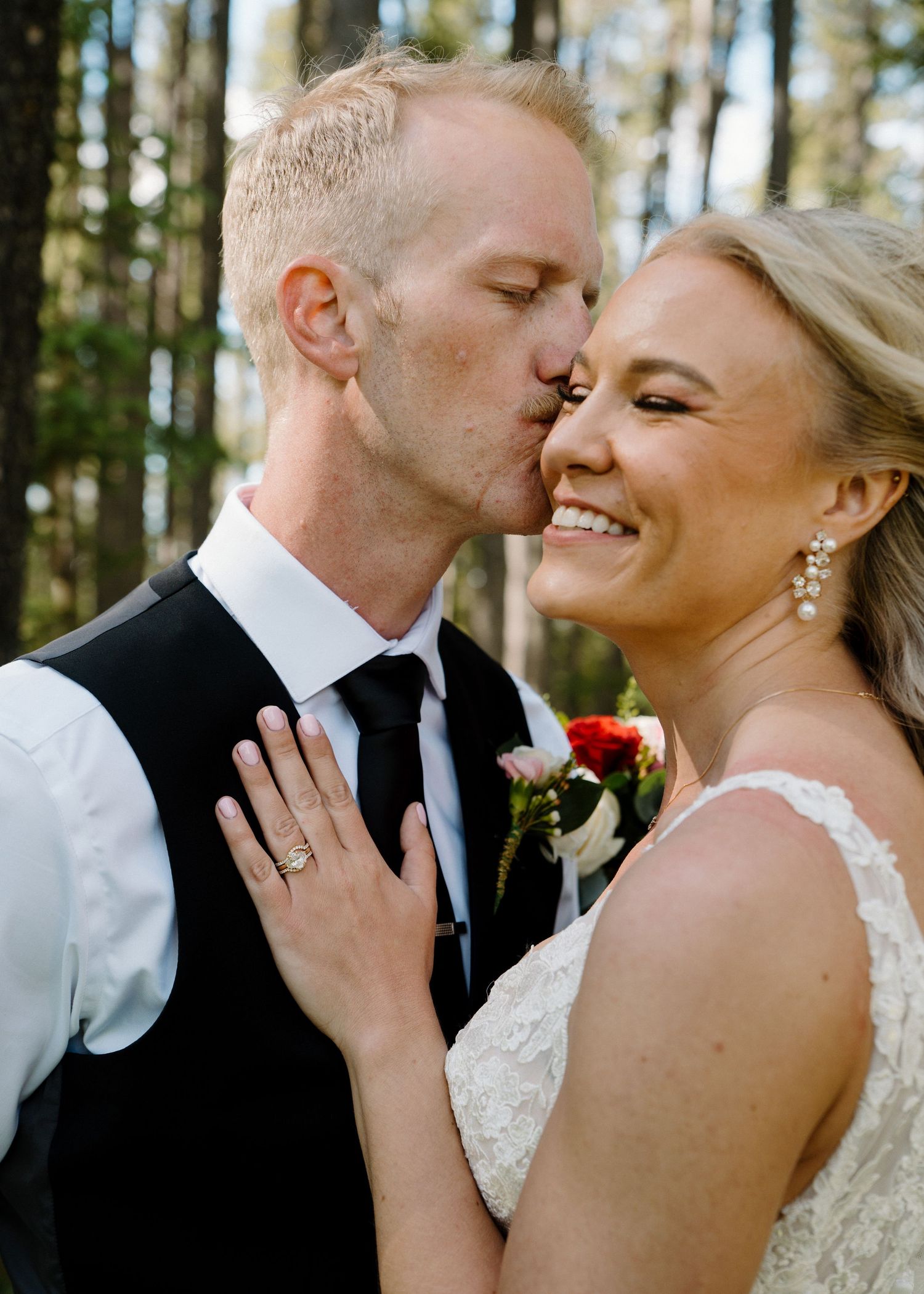 Groom in black vest and tie kisses bride on cheek as she smiles, wearing lace wedding dress and floral bouquet.