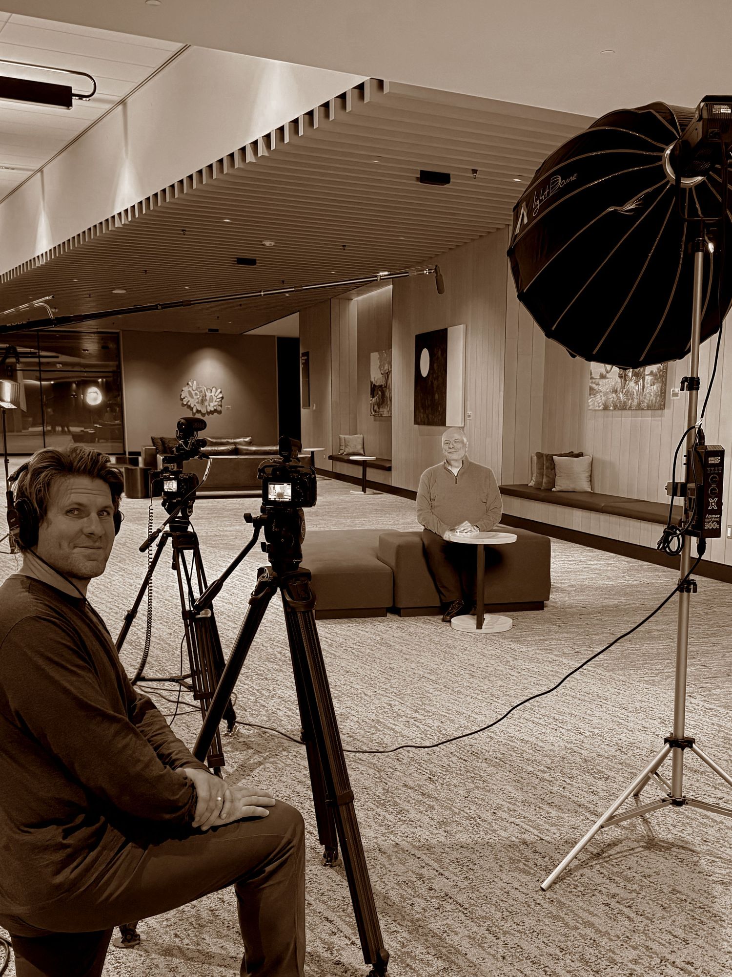 Sepia-toned behind-the-scenes photo of a videographer filming an interview in a stylish contemporary corporate space.