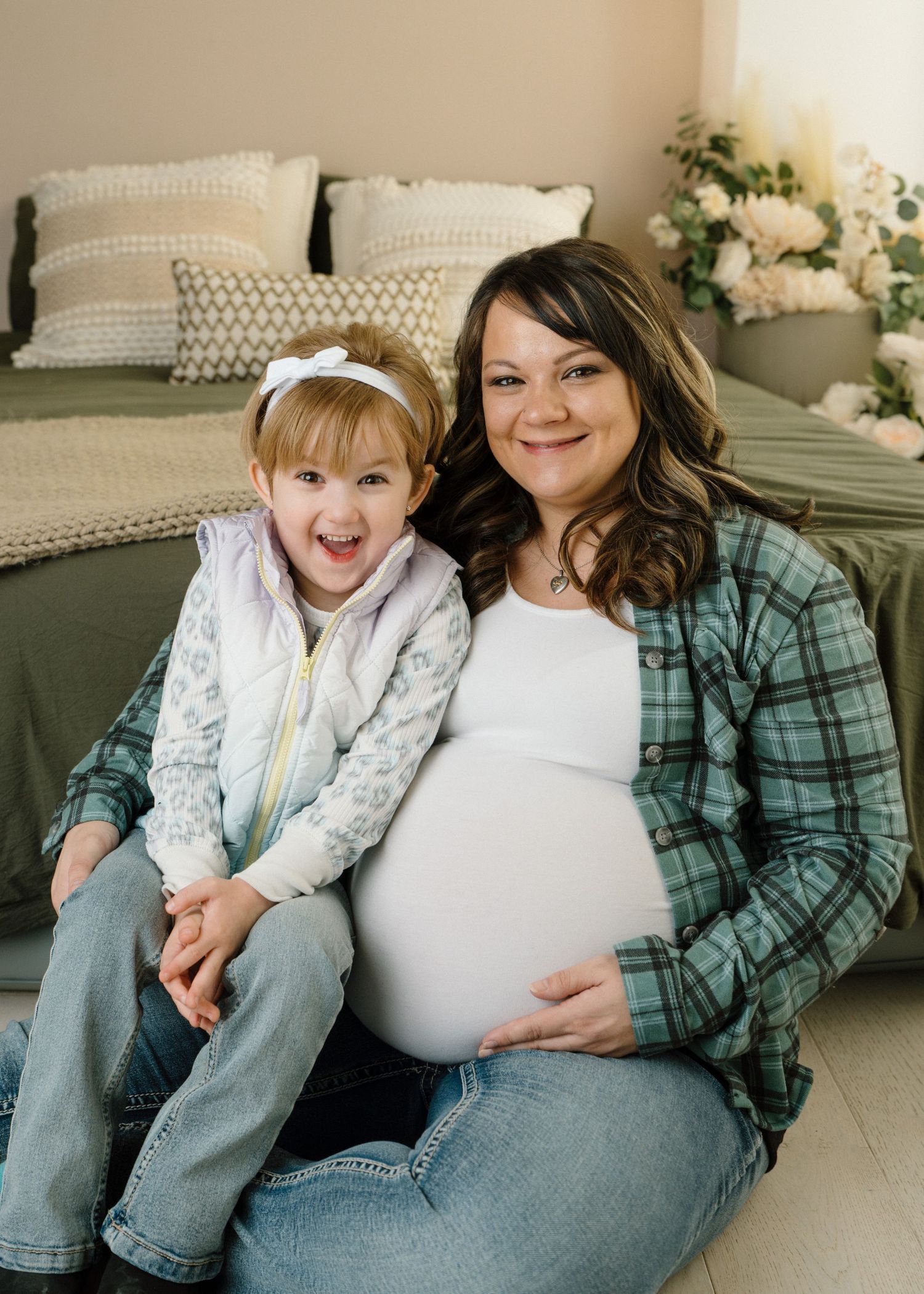 Pregnant mother in plaid shirt sitting on floor with excited toddler daughter in purple vest and bow.
