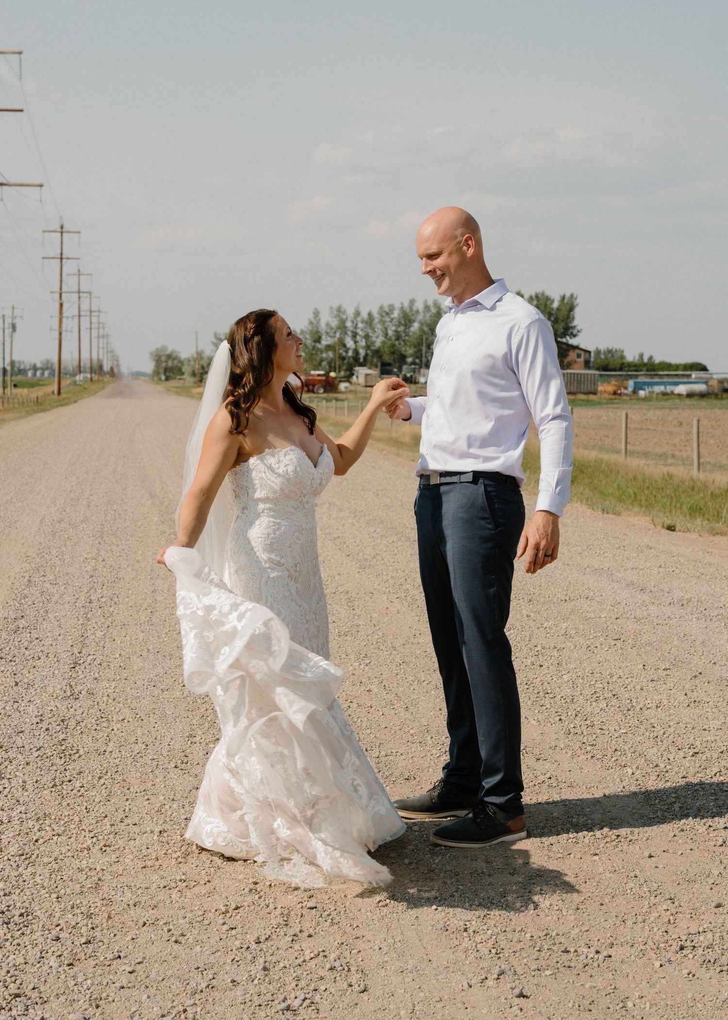 Bride in lace gown twirls as groom smiles on a rural dirt road during wedding portraits.