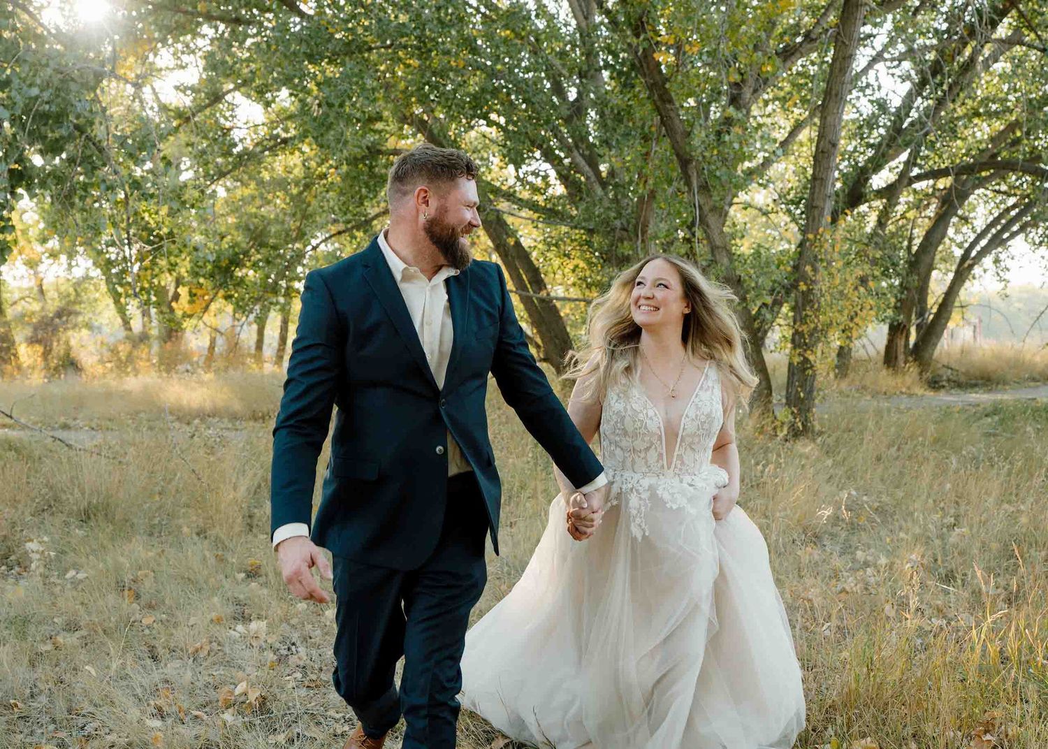 A joyful couple walks hand-in-hand through a sunlit meadow, bride in lace gown and groom in navy suit in Medicine Hat Alberta