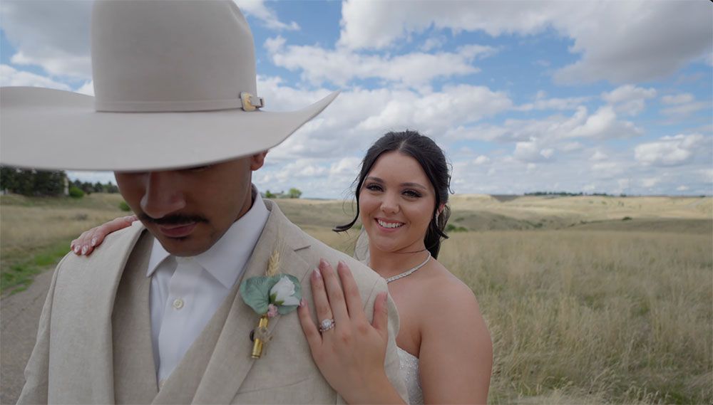 Bride and groom in western attire pose in golden field, she displays ring while he wears white cowboy hat.