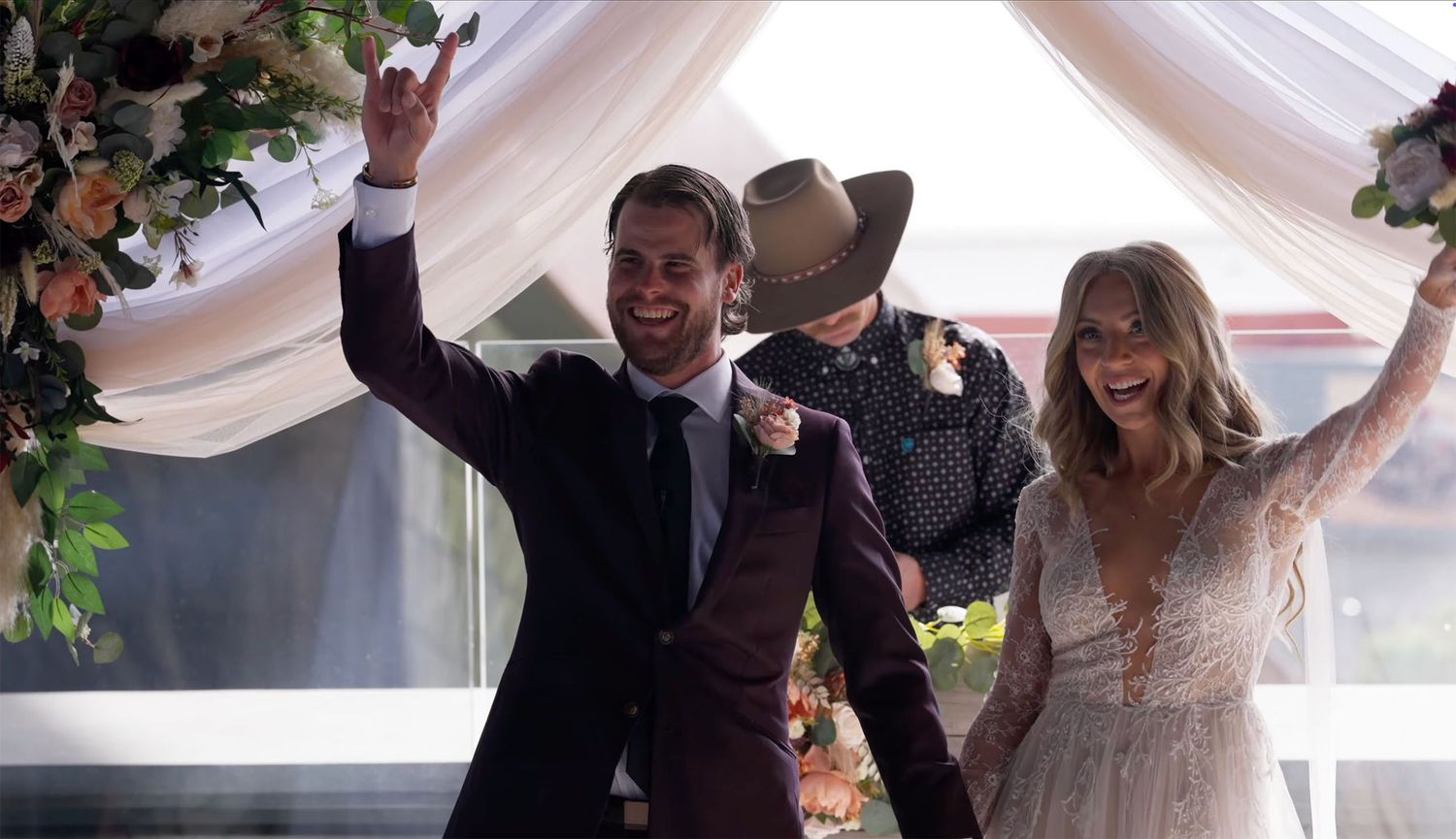 Joyful couple celebrates wedding ceremony under floral arch, groom in burgundy suit, bride in lace gown, raising hands.