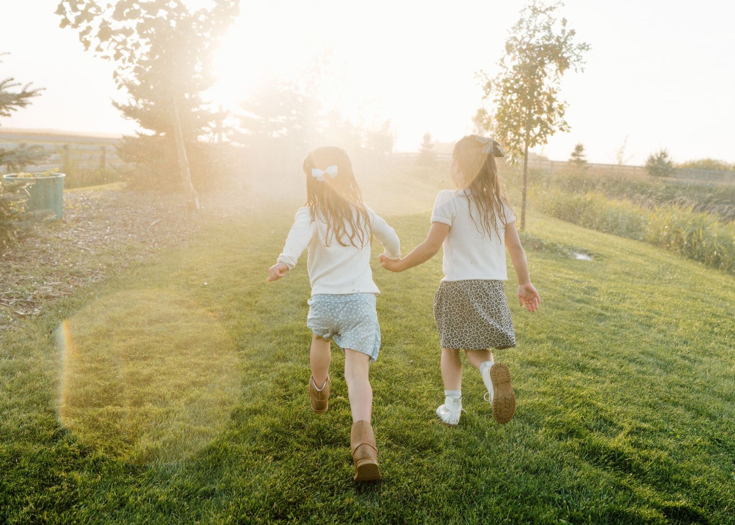 Two young girls holding hands running through a sunny green field at golden hour, viewed from behind.