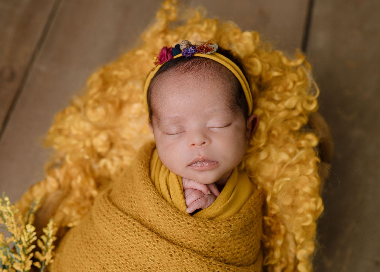 Sleeping newborn baby swaddled in mustard yellow wrap with floral headband on fluffy yellow backdrop.