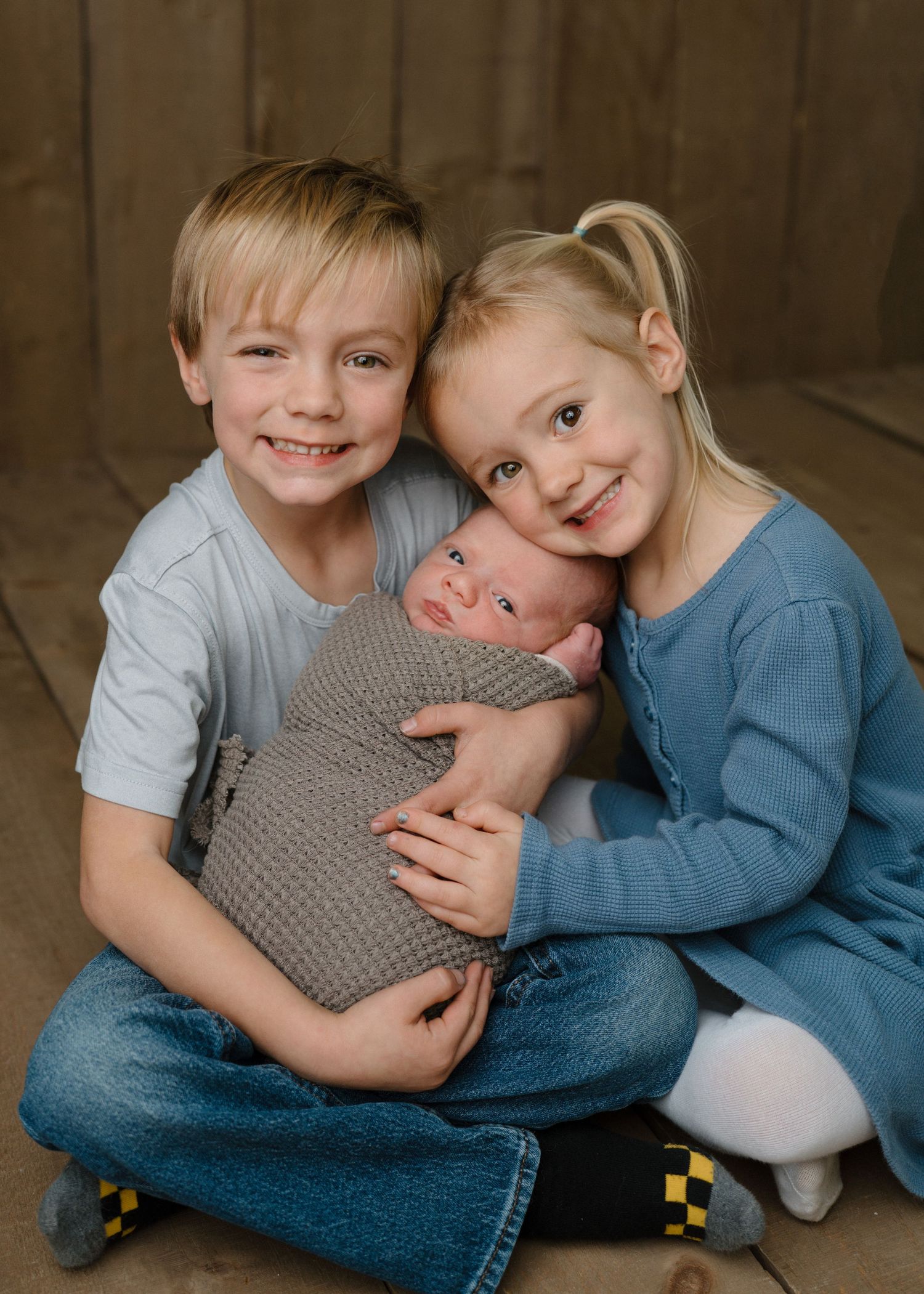 Two smiling siblings in blue and white holding a swaddled newborn baby on a wooden floor.