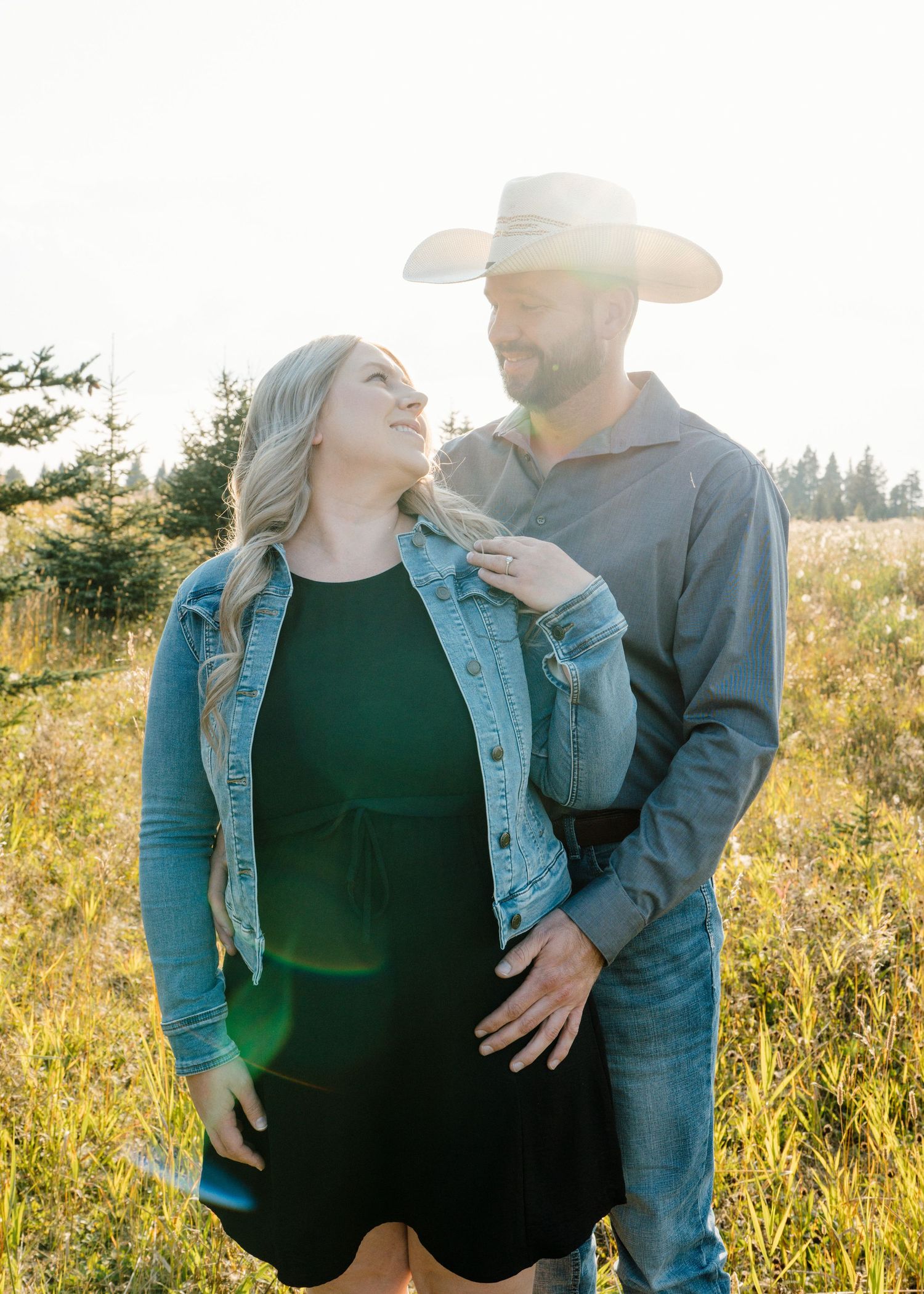 Couple gazes at each other in sunny field, woman in denim jacket and black dress.