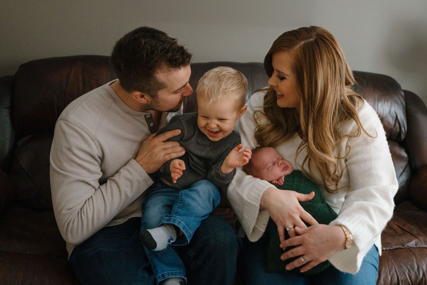 Happy young family of four sitting on brown leather couch with toddler and newborn baby.