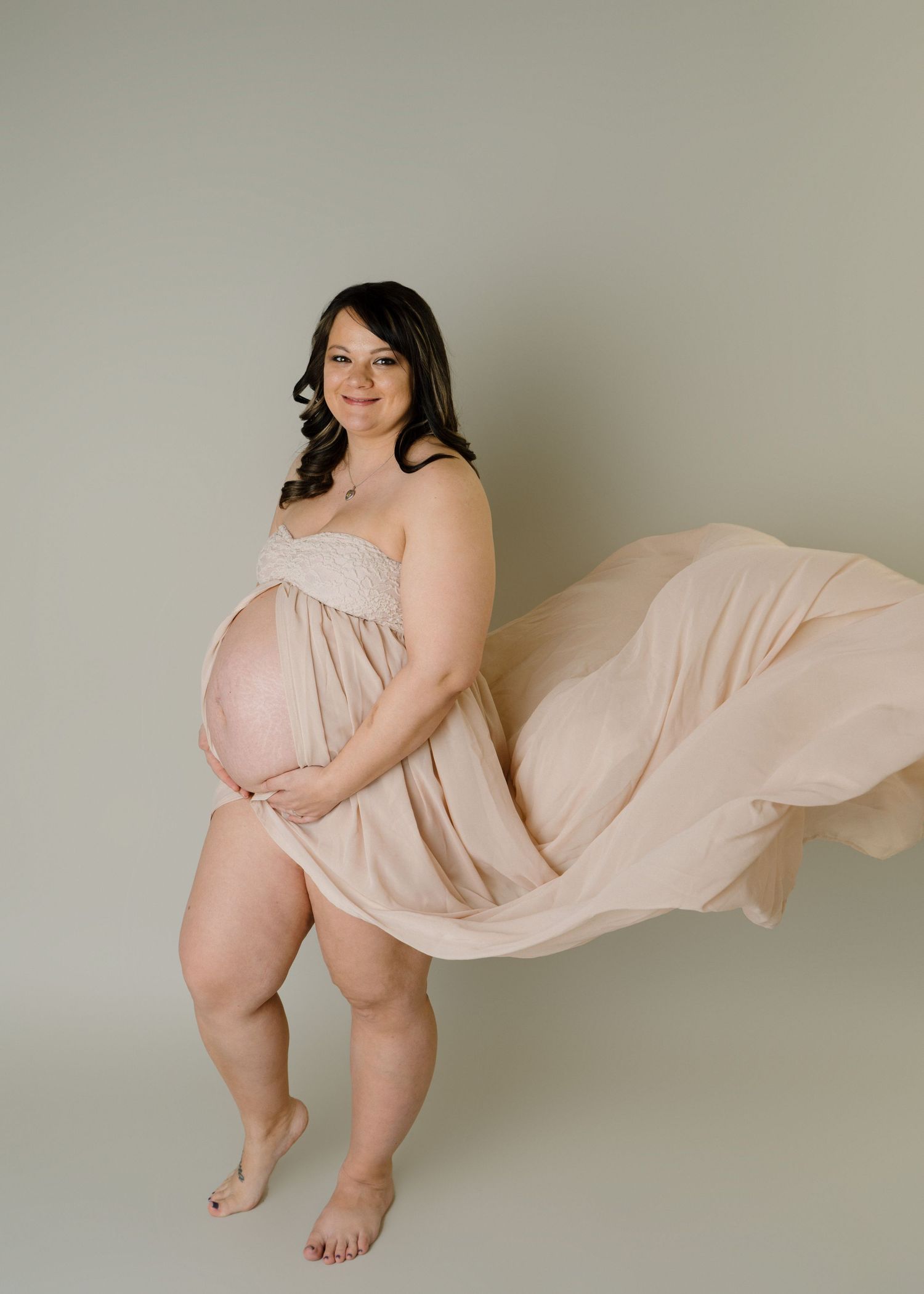 Pregnant woman in flowing beige fabric dress with billowing skirt posing barefoot in studio portrait.