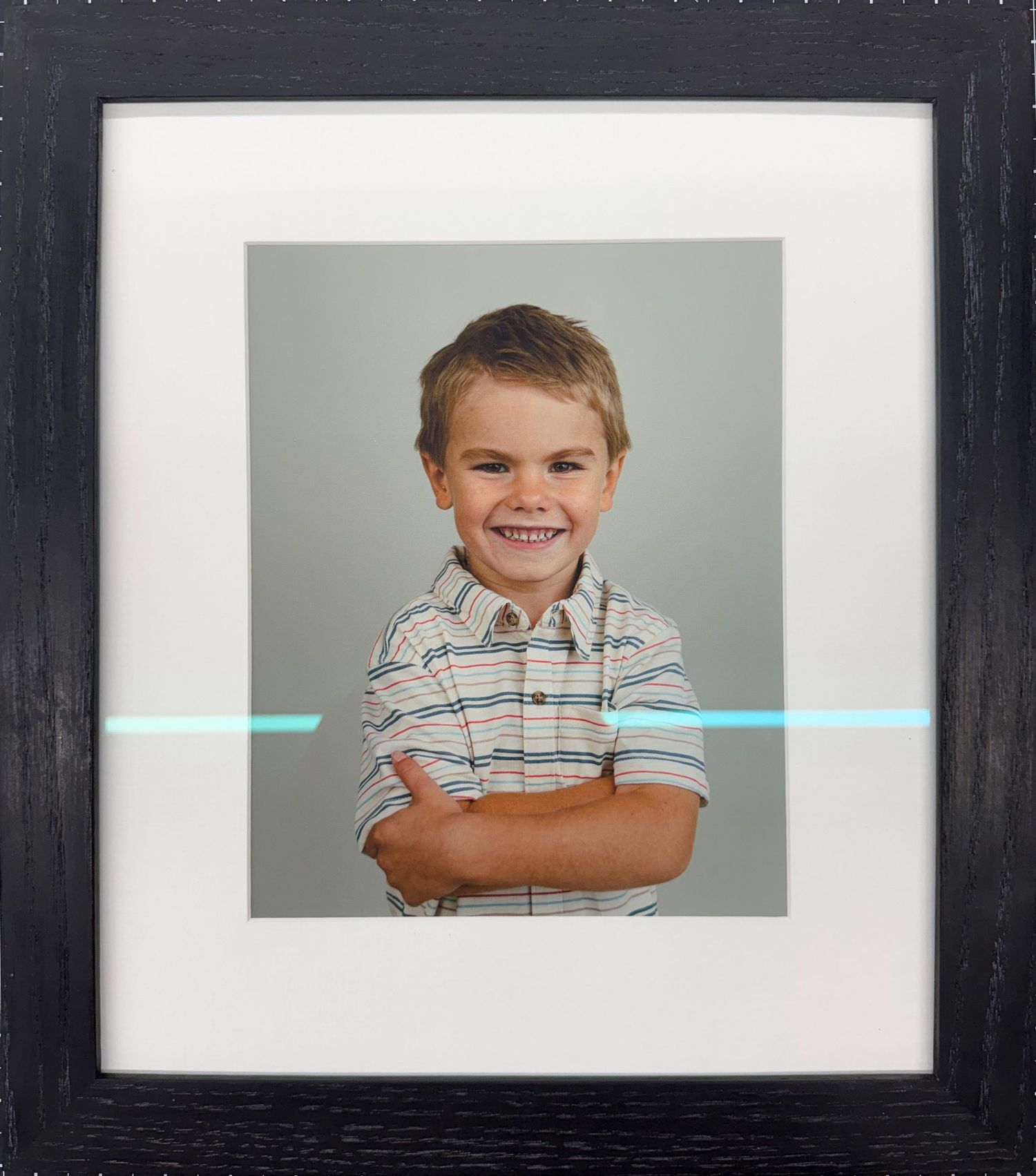 Smiling young boy with crossed arms in a striped polo shirt, displayed in a black picture frame with white matting.