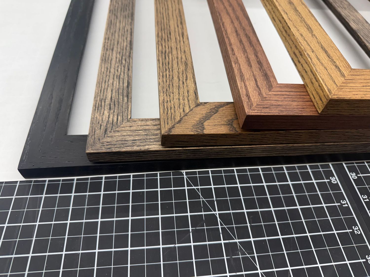 Multiple wood picture frame samples in various stains displayed on a cutting mat, showcasing dark, medium, and light wood finishes.