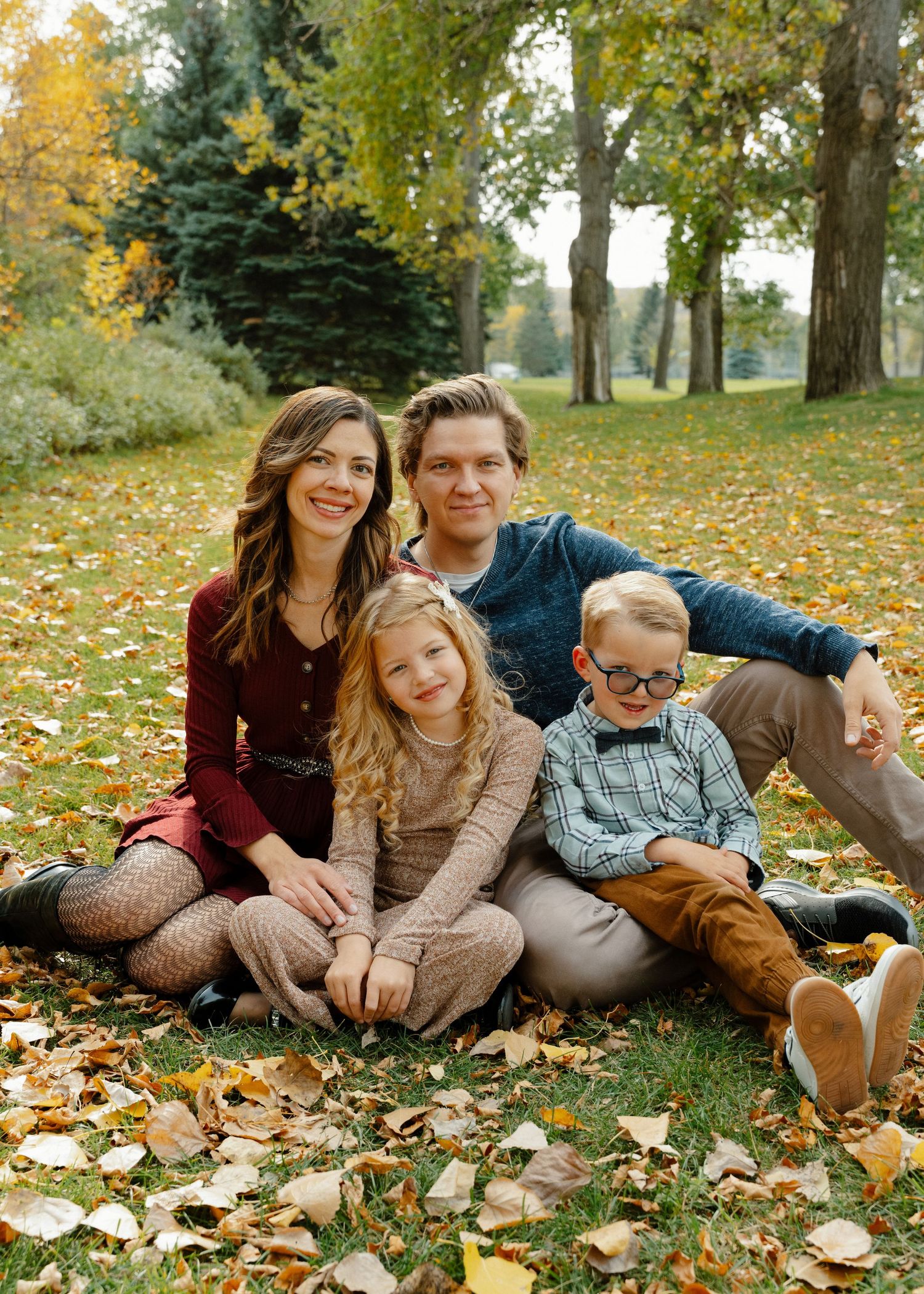 Family of four sitting together on fall leaves in a sunlit park setting.