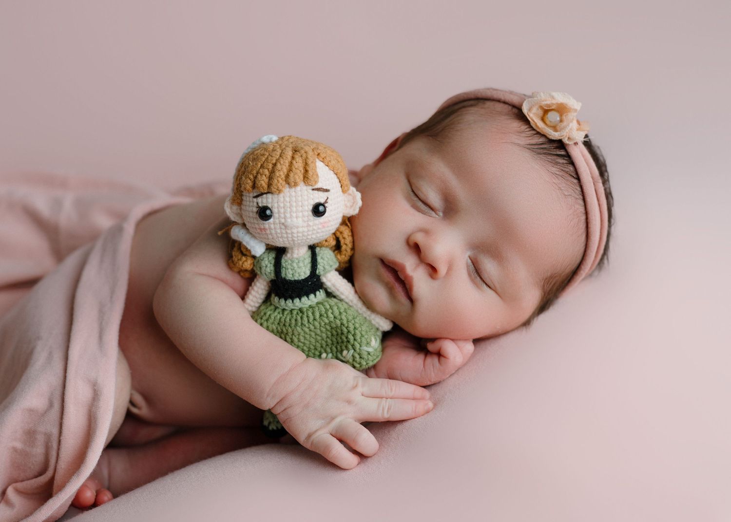 Sleeping newborn baby girl holding a crocheted Anna doll, wearing a pink headband with flower.