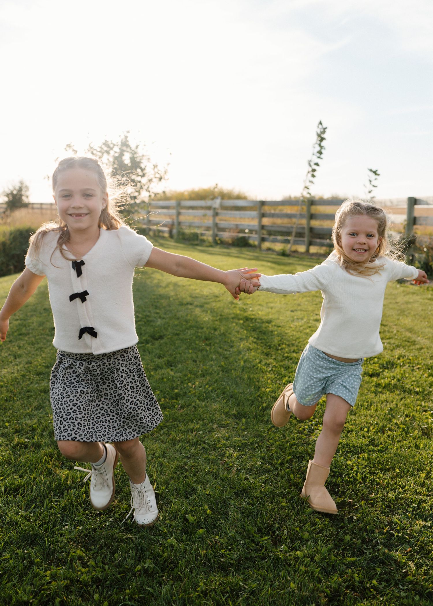 Two smiling girls in white tops hold hands running joyfully across a green lawn at sunset.