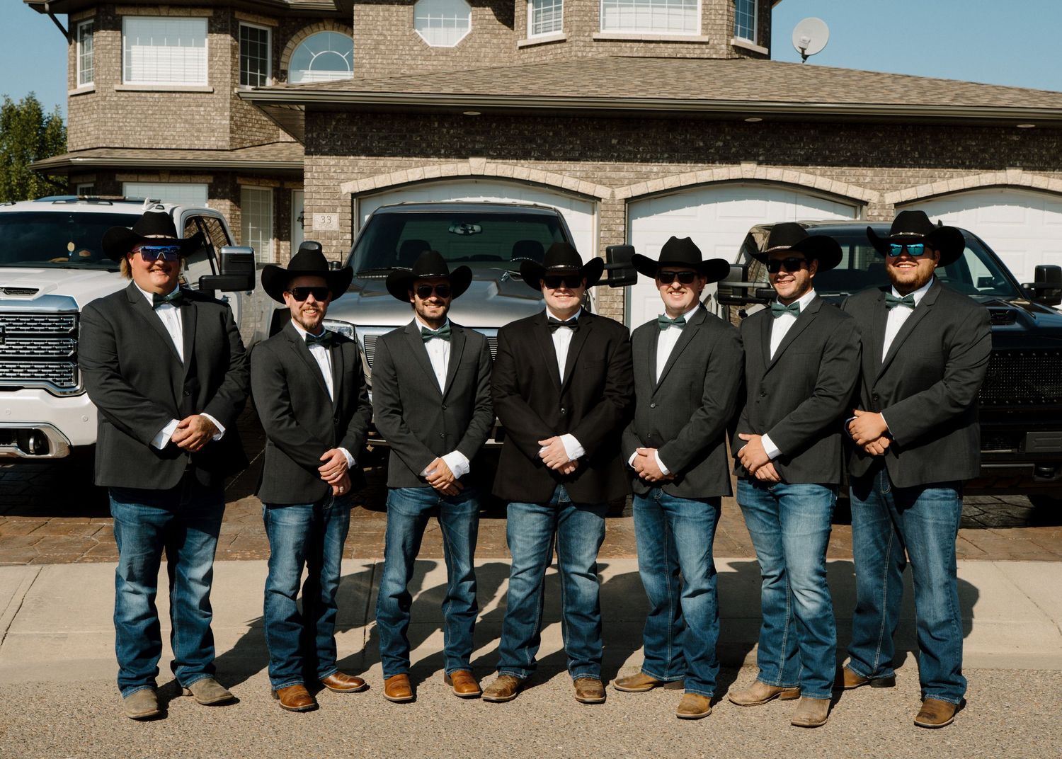 Seven groomsmen in black tuxedos, cowboy hats, and jeans posing in front of trucks.