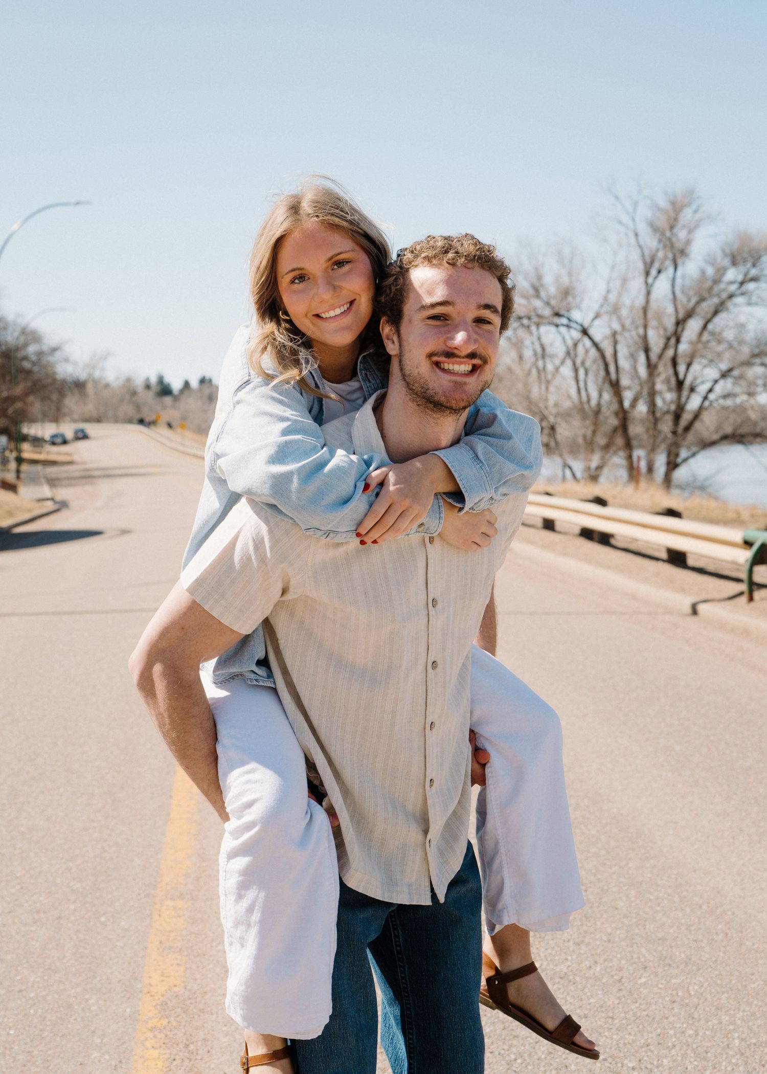 Young couple smiling on a road, woman piggyback riding on mans back on sunny day.