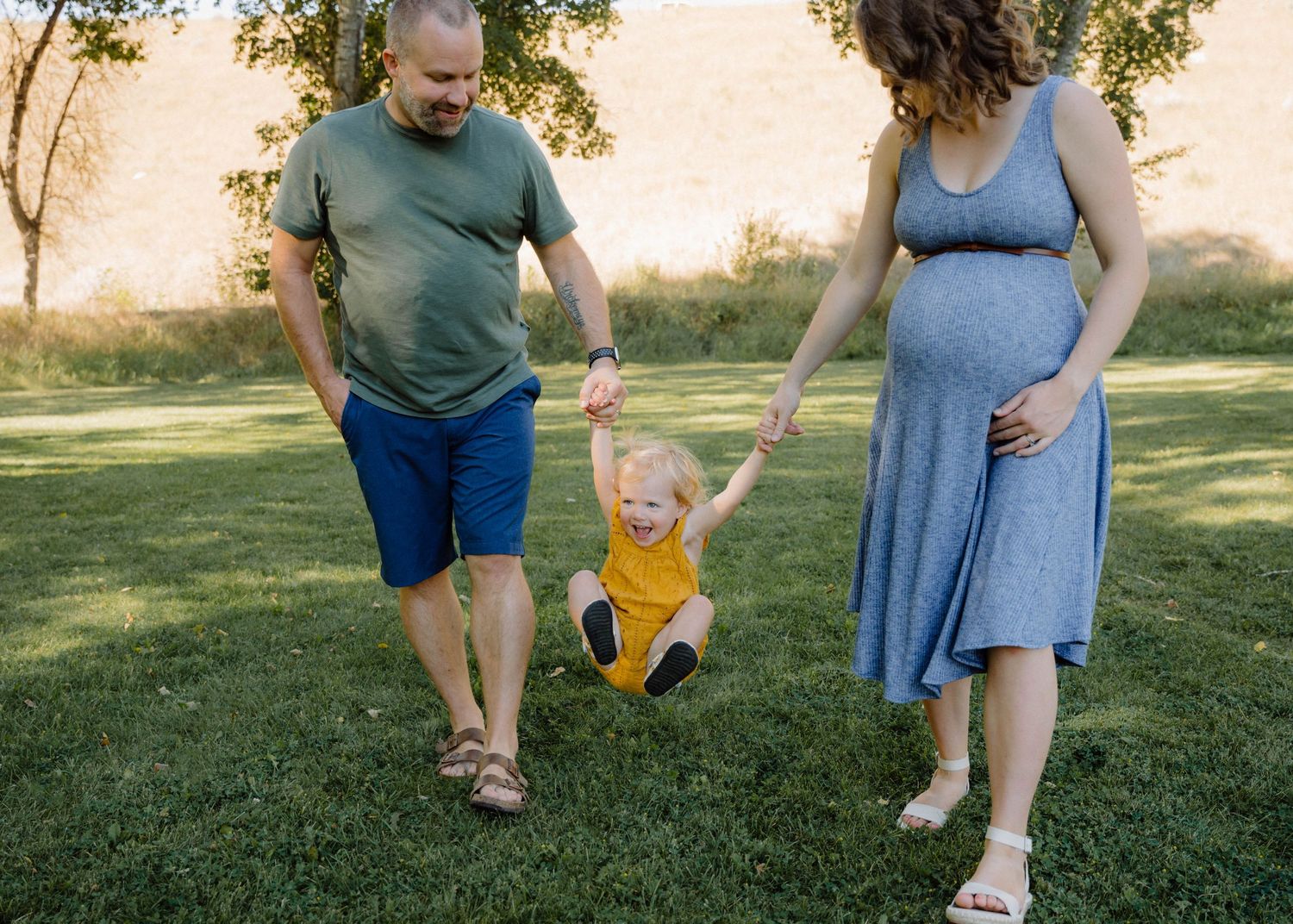 Pregnant couple in park swinging their laughing toddler daughter in yellow romper between them on green grass.
