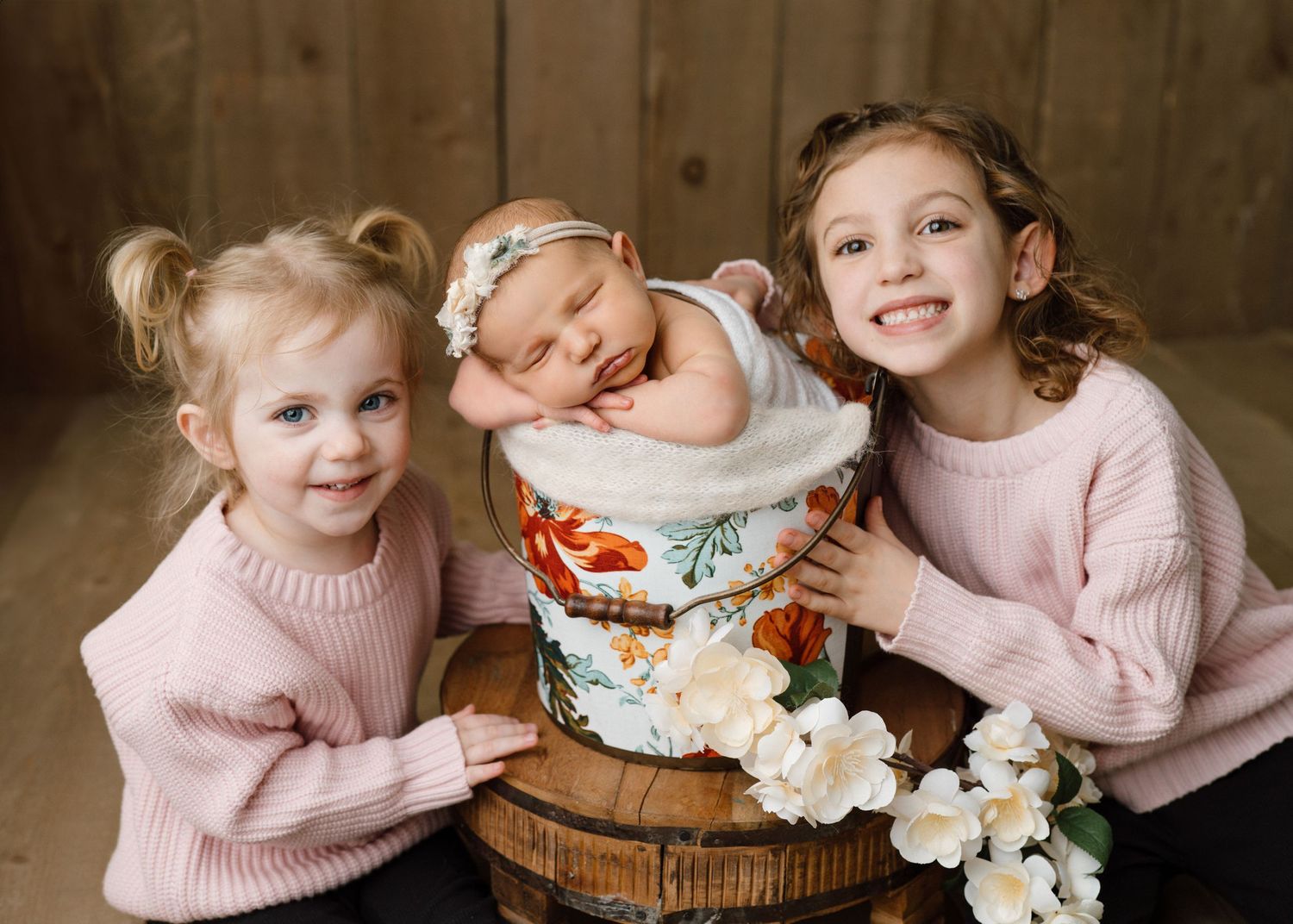 Two young girls in pink sweaters posing with sleeping newborn in floral bucket with white roses.