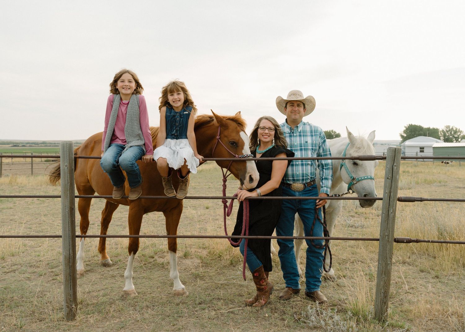 Two girls sitting on horses at fence with parents standing alongside two horses on ranch.