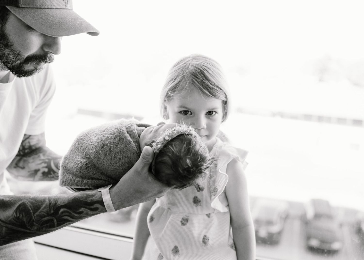 Black and white photo of tattooed dad holding newborn while young daughter looks on.
