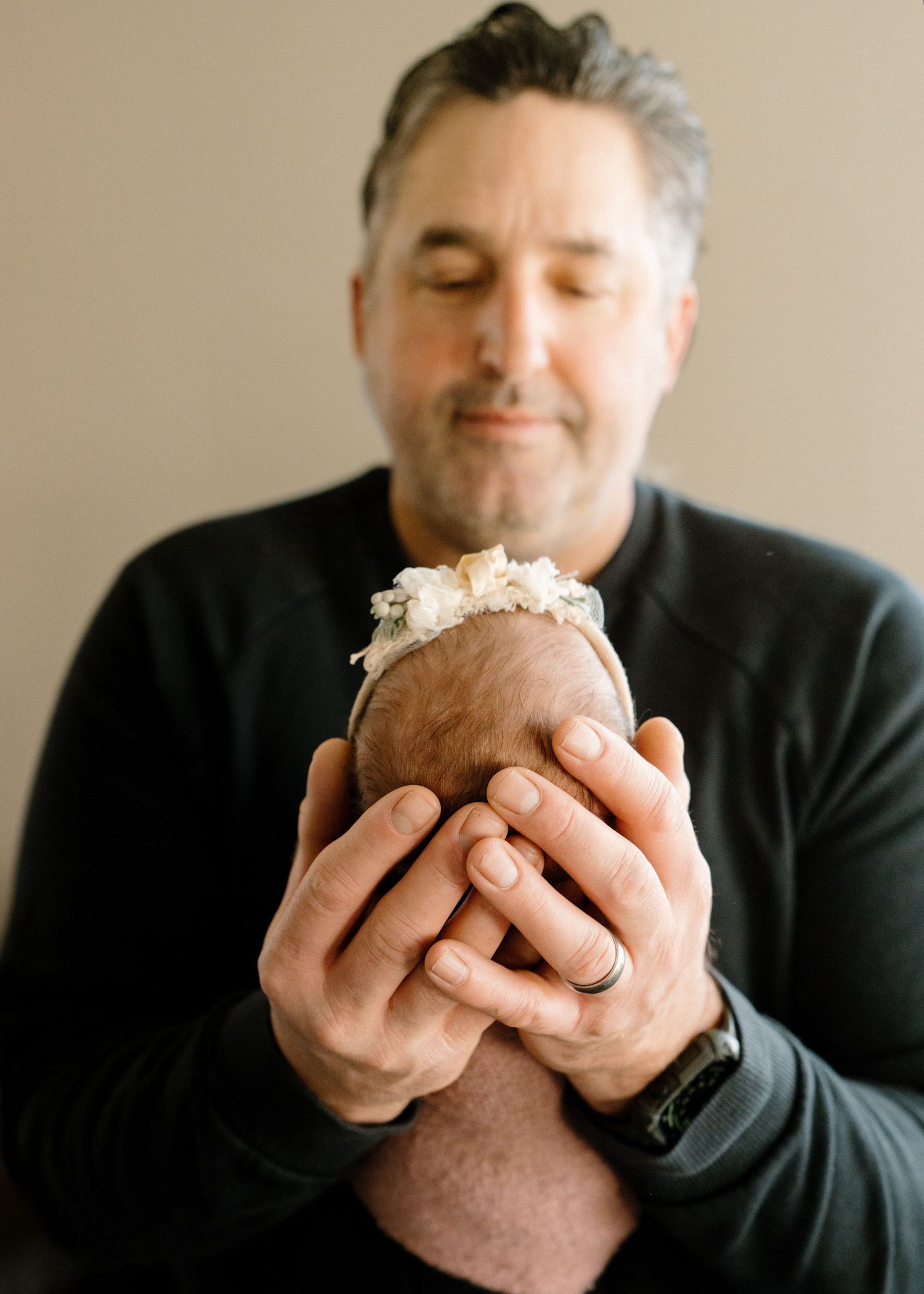 Father in black shirt gently cradling tiny newborn baby with floral headband in his hands.