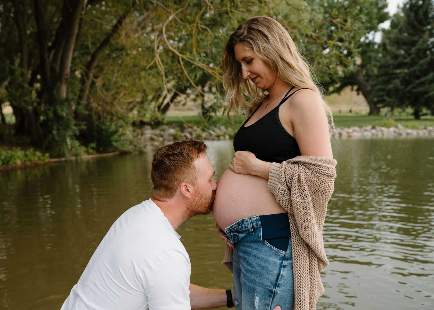 Man kneeling to kiss pregnant woman's belly while standing in a peaceful pond.