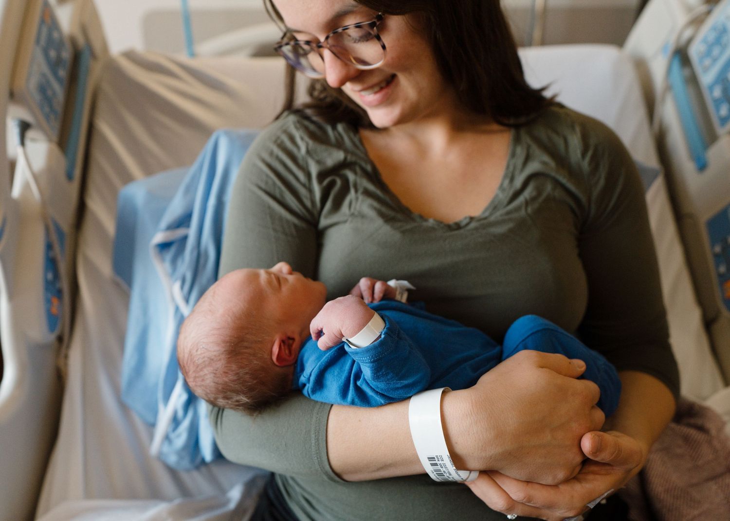 Smiling new mother in hospital gown holding newborn baby boy wrapped in blue blanket after birth.