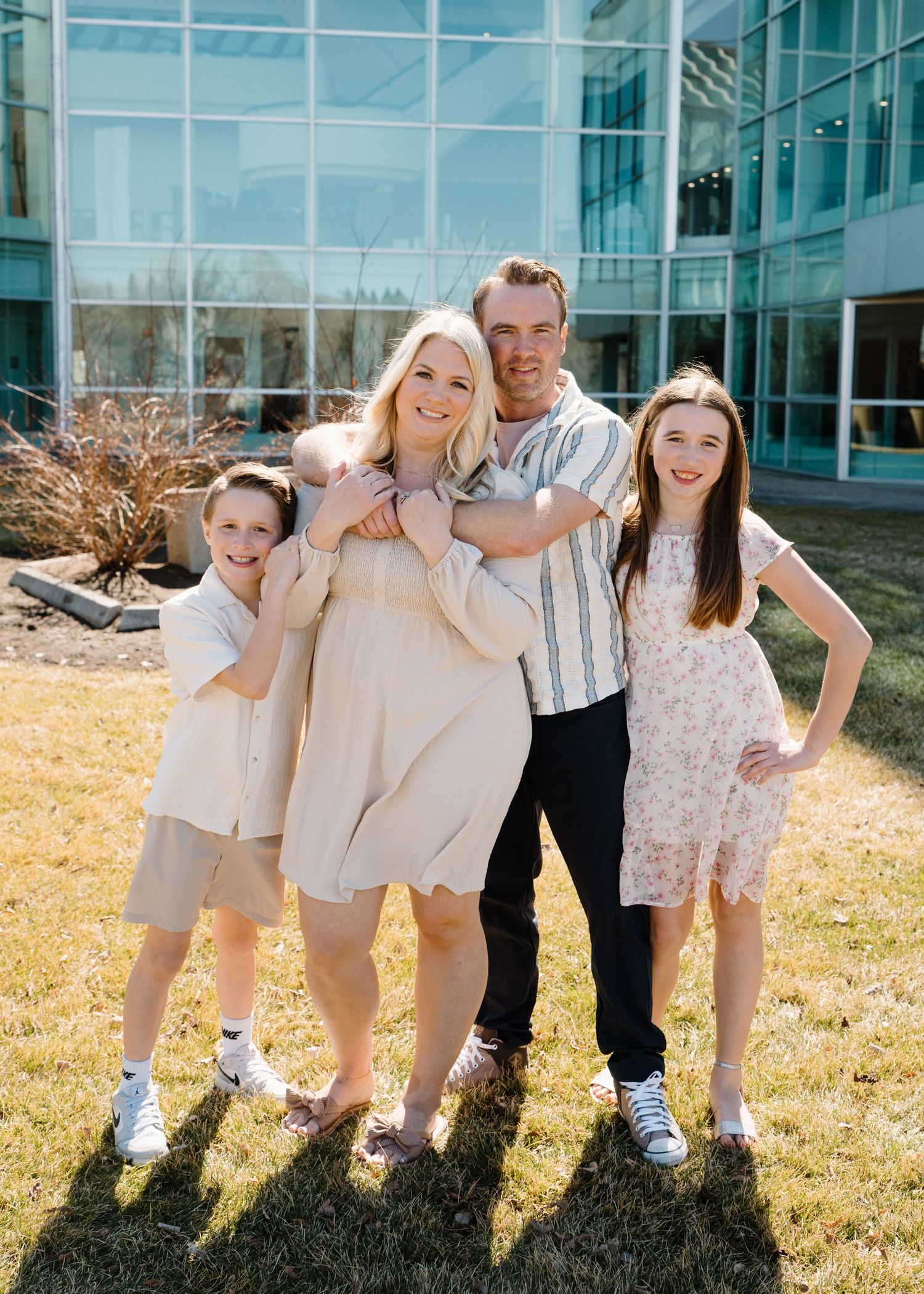 Happy family of four posing outdoors in front of a modern glass building on sunny day.