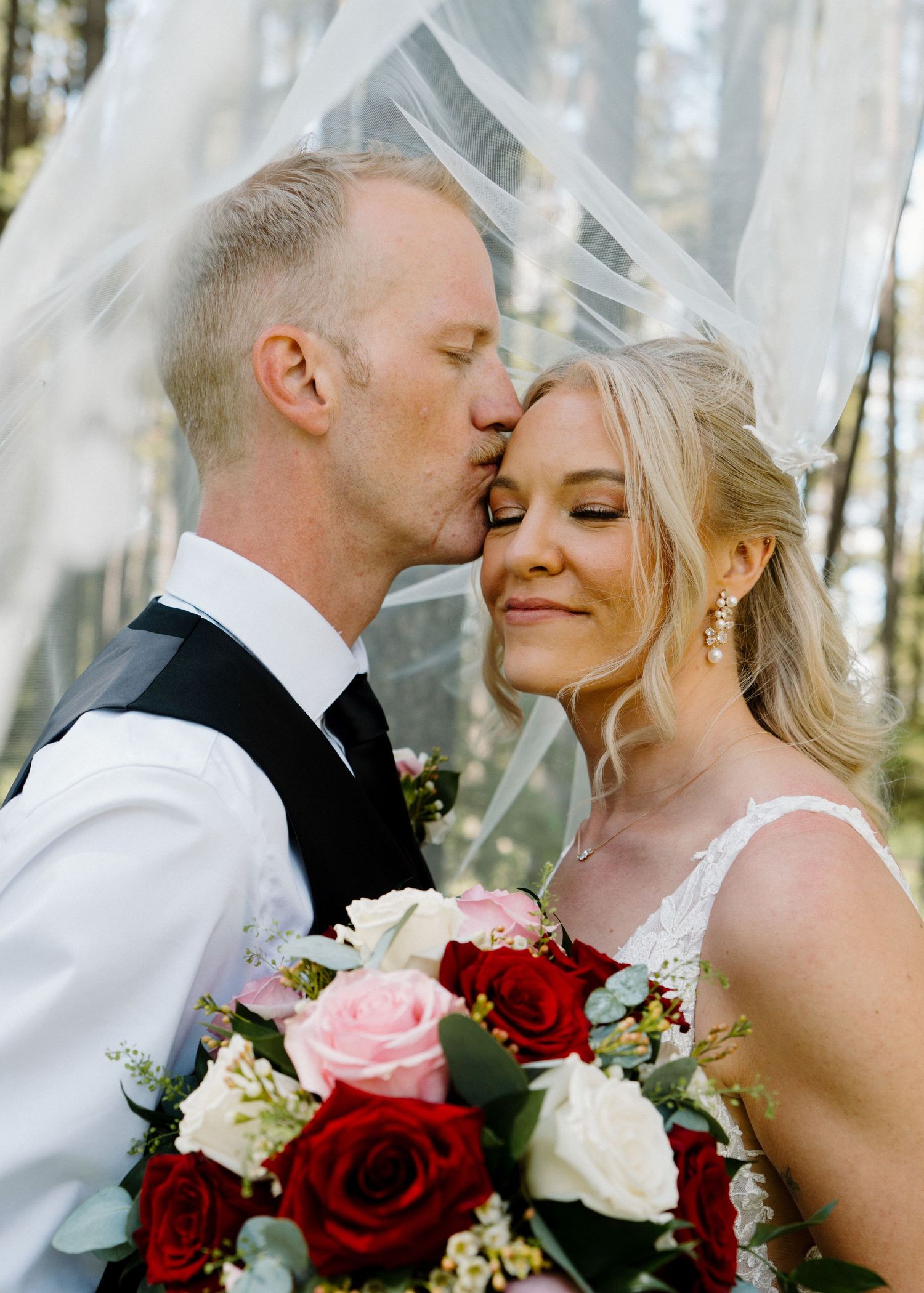 Groom kissing bride on cheek under veil, bride holding red and pink rose bouquet, outdoor wedding photo.
