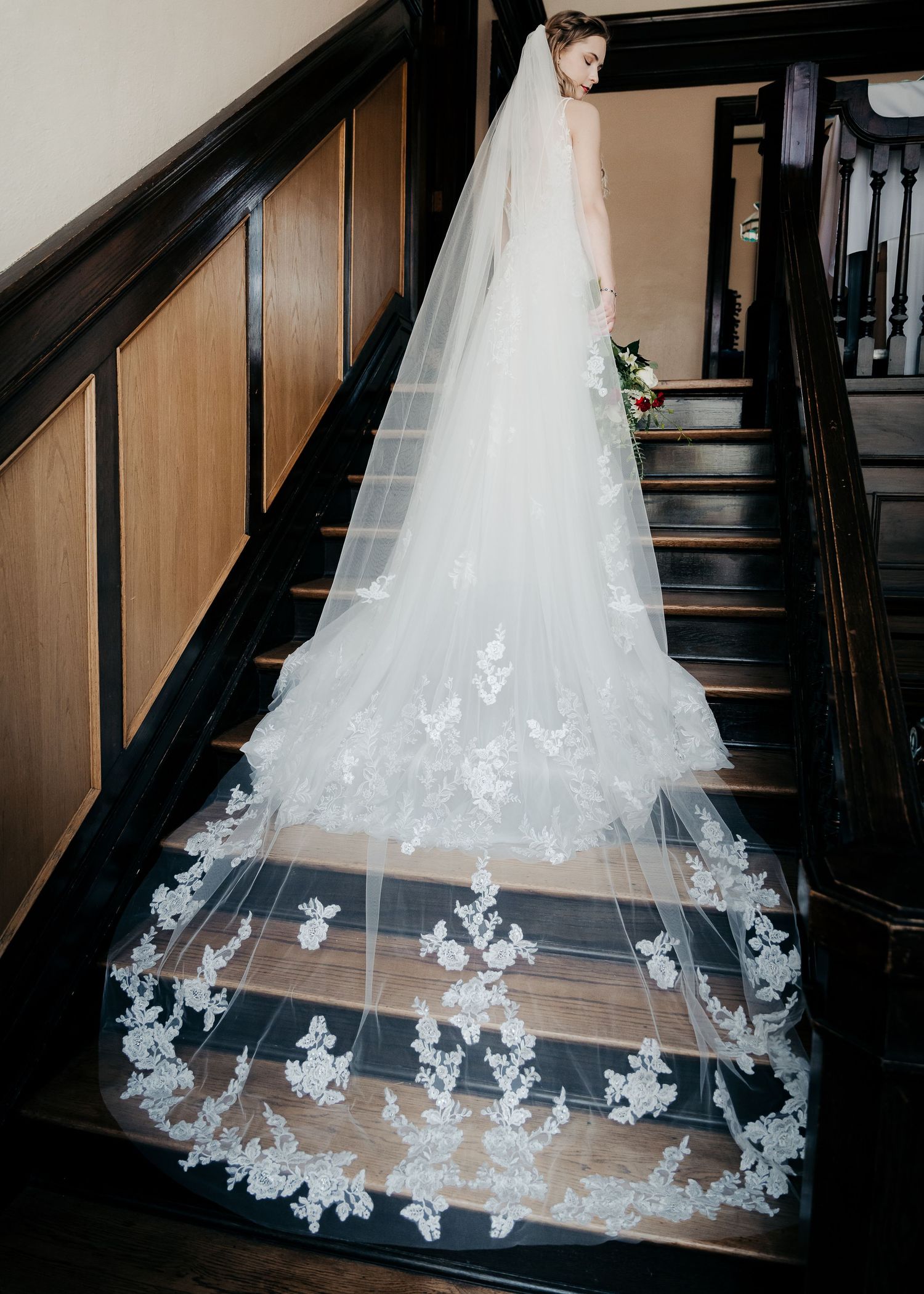 Bride in elegant gown with long lace cathedral veil trailing down dark wood staircase.