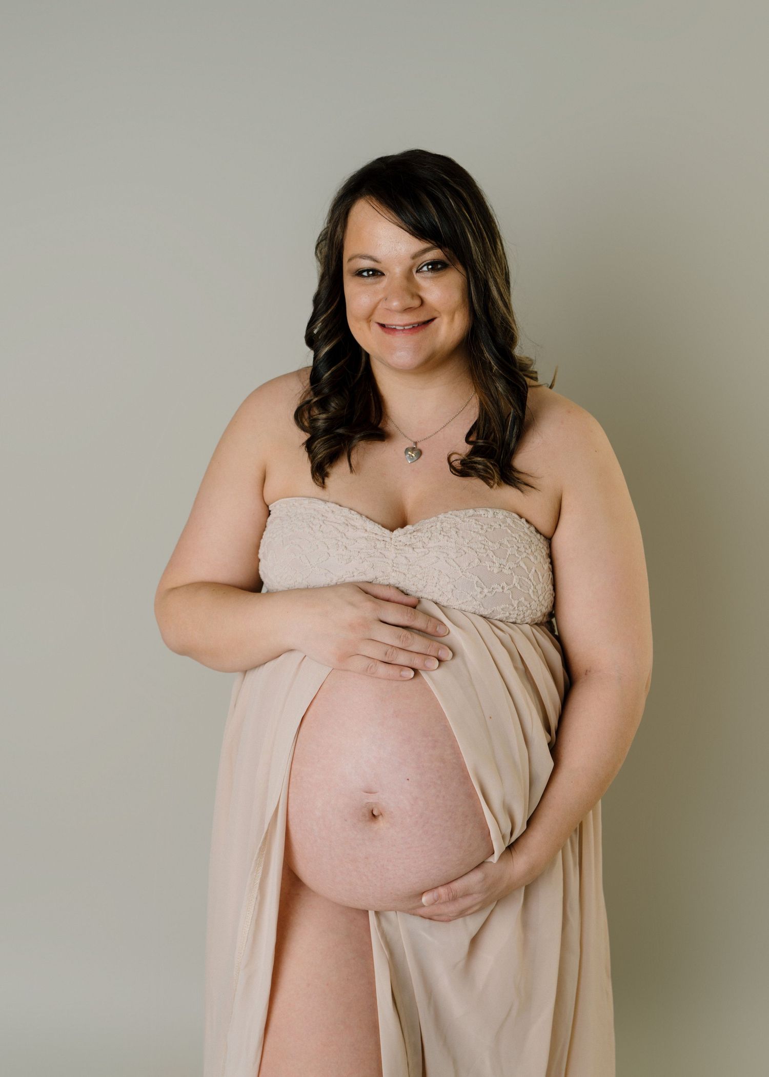 Smiling pregnant woman in flowing beige strapless gown cradling her bare baby bump in a studio portrait.