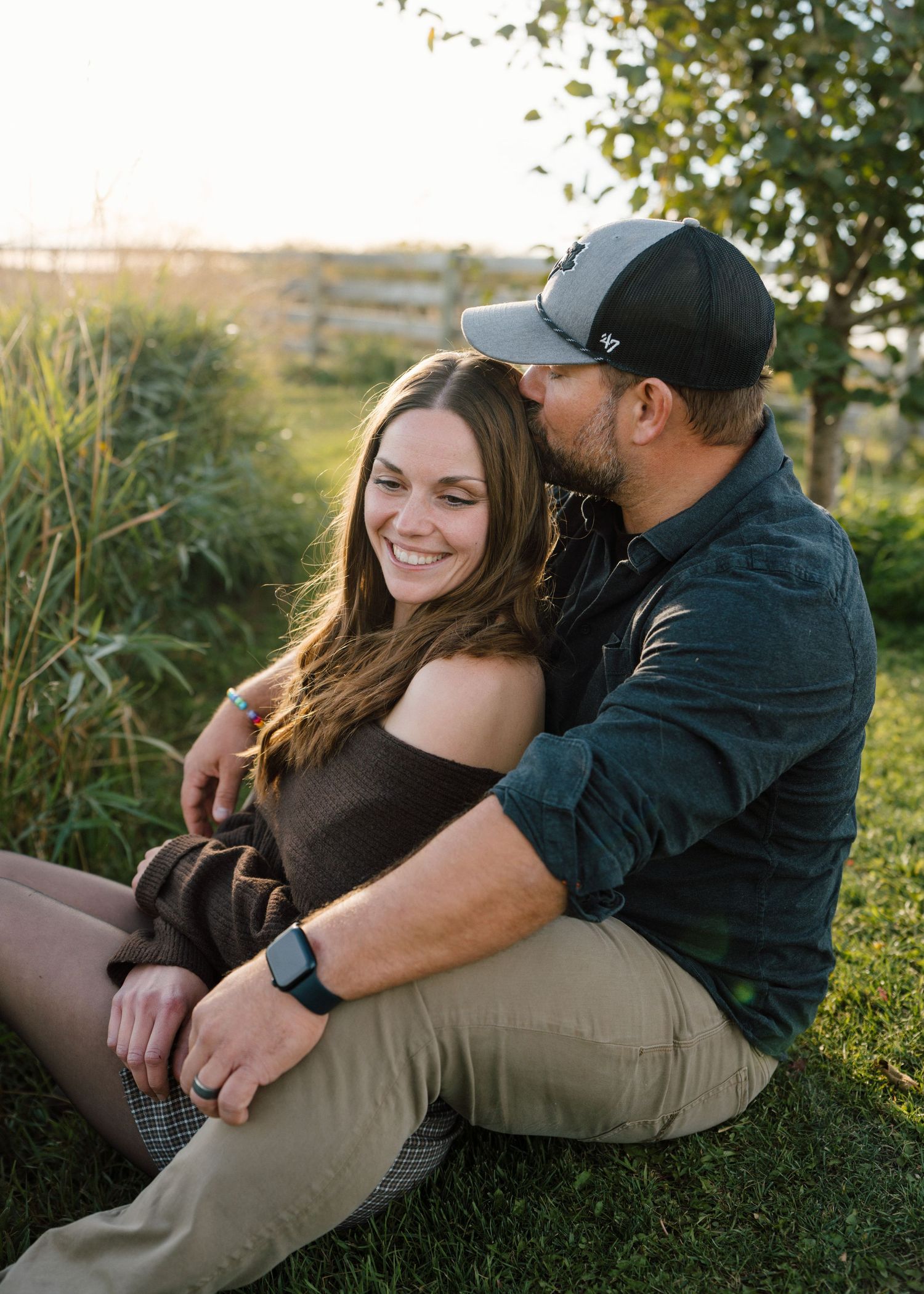 A couple sits together in grass at golden hour, woman smiling as man kisses her cheek.