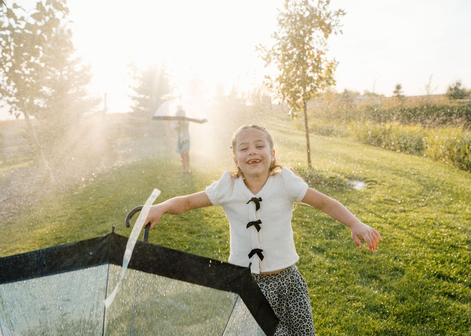 Young girl with arms outstretched plays joyfully near a sprinkler in a sunny backyard garden.
