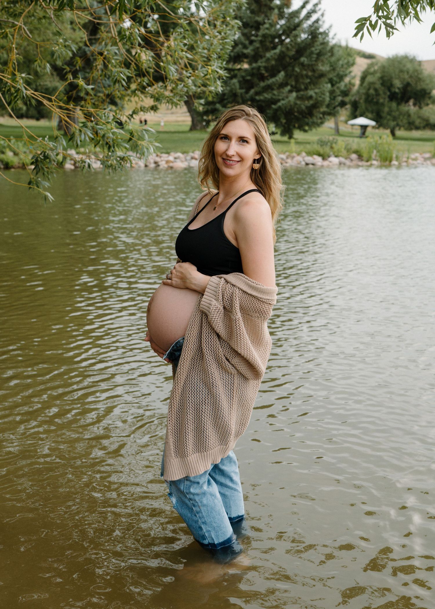 Pregnant woman in black top and jeans with beige cardigan standing in a serene pond.