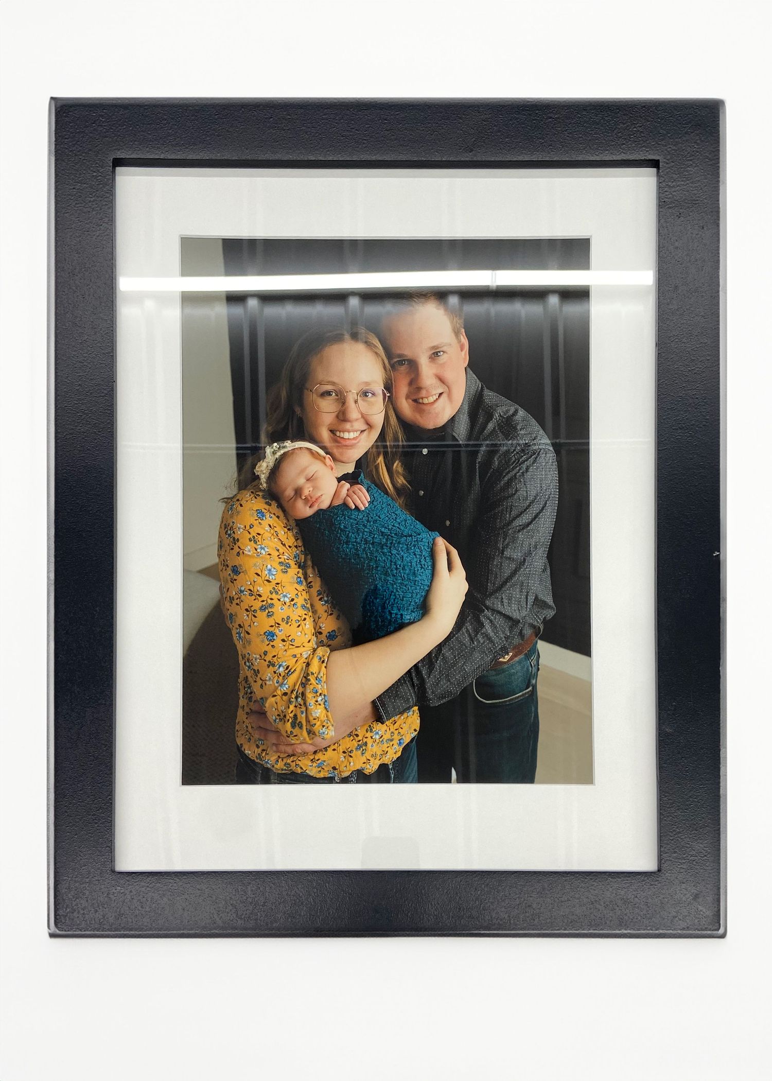 Black framed matted family portrait with newborn baby displayed on white wall.