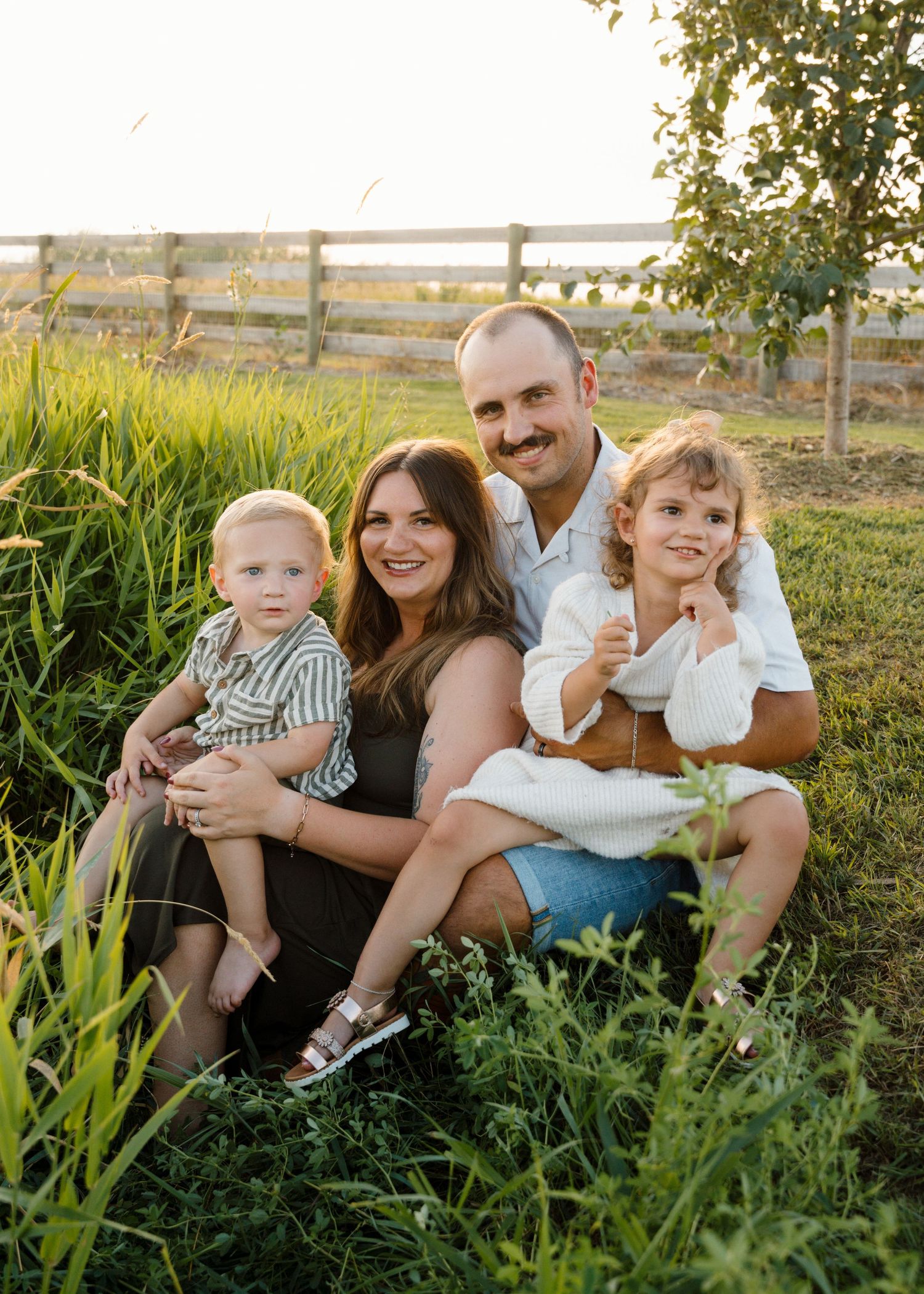 Family of four sitting in lush green grass near a wooden fence at sunset.