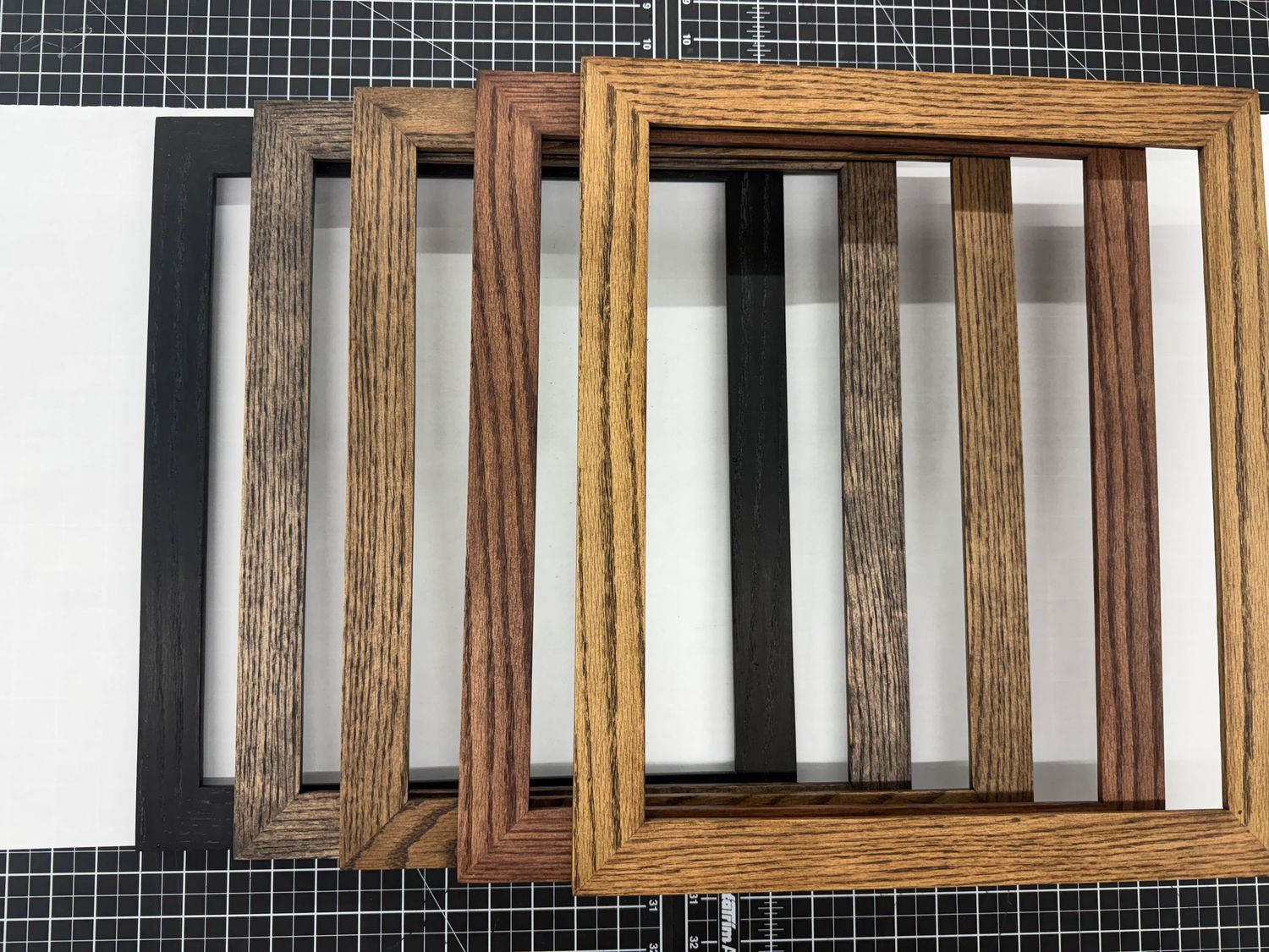 Five wooden picture frames in various shades from black to light oak arranged in a stacked display on a dark grid surface.