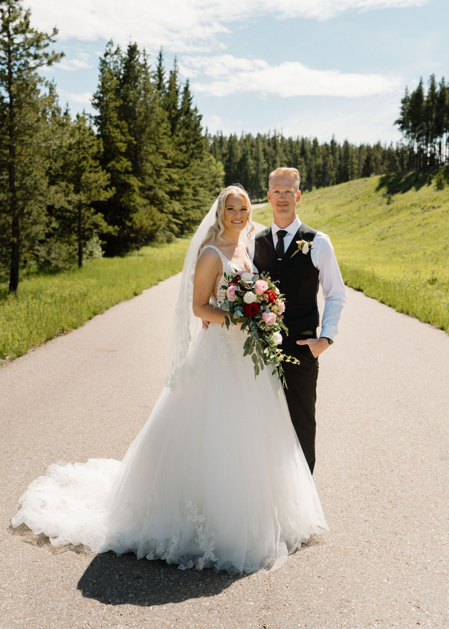 Bride in white gown with veil holding bouquet stands with groom in black suit on scenic mountain road surrounded by trees.
