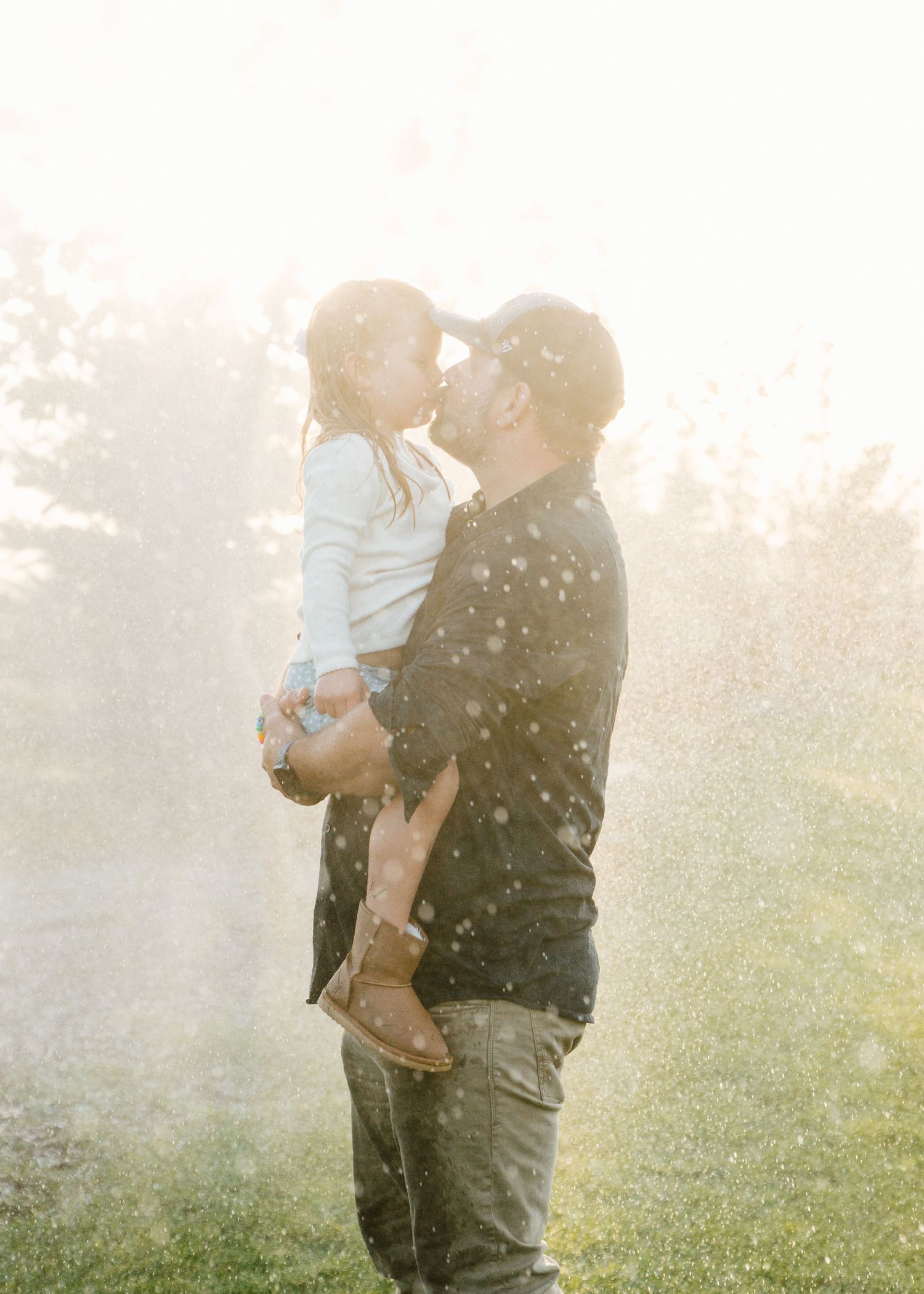 A father holds his young child for a tender kiss outdoors in golden sunlight with water droplets sparkling around them.