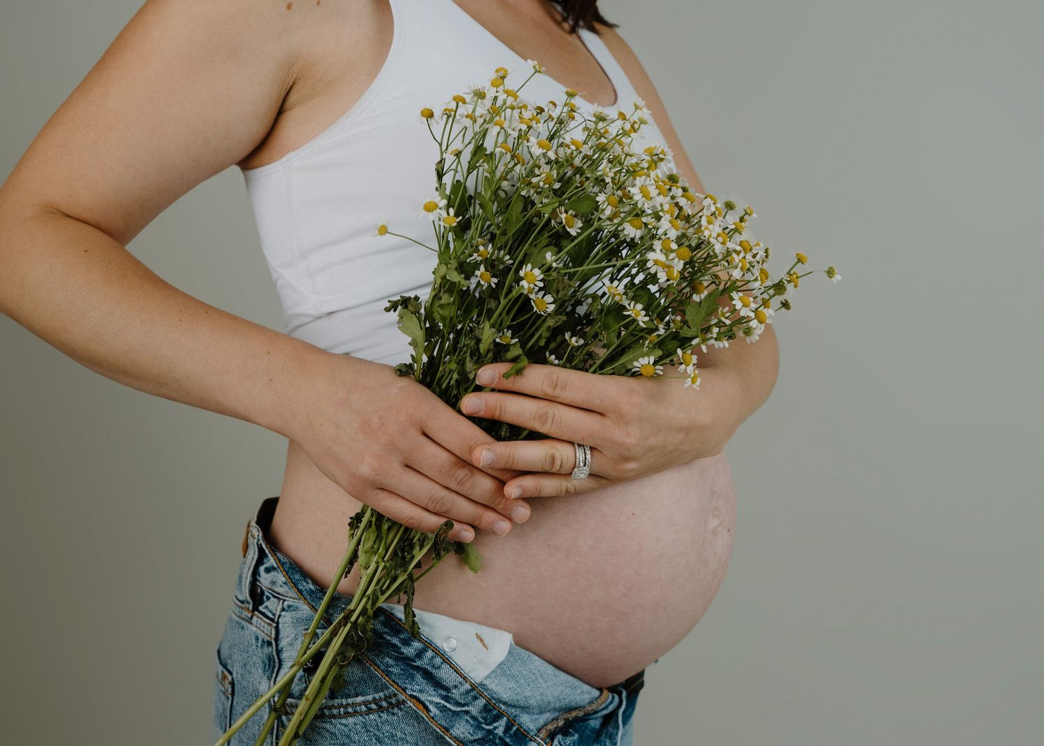 Pregnant woman in white crop top and jeans holding a bouquet of white wildflowers against a gray background.