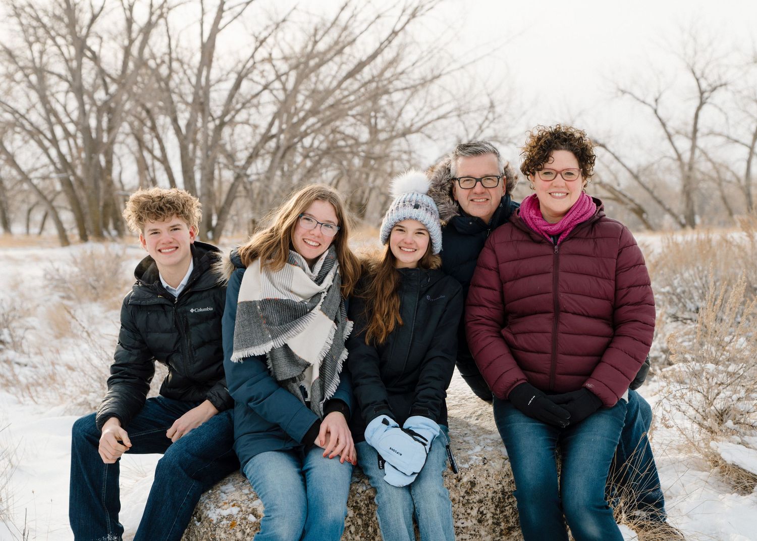 Family of five smiling together outdoors in a snowy winter landscape, wearing cozy jackets and scarves.