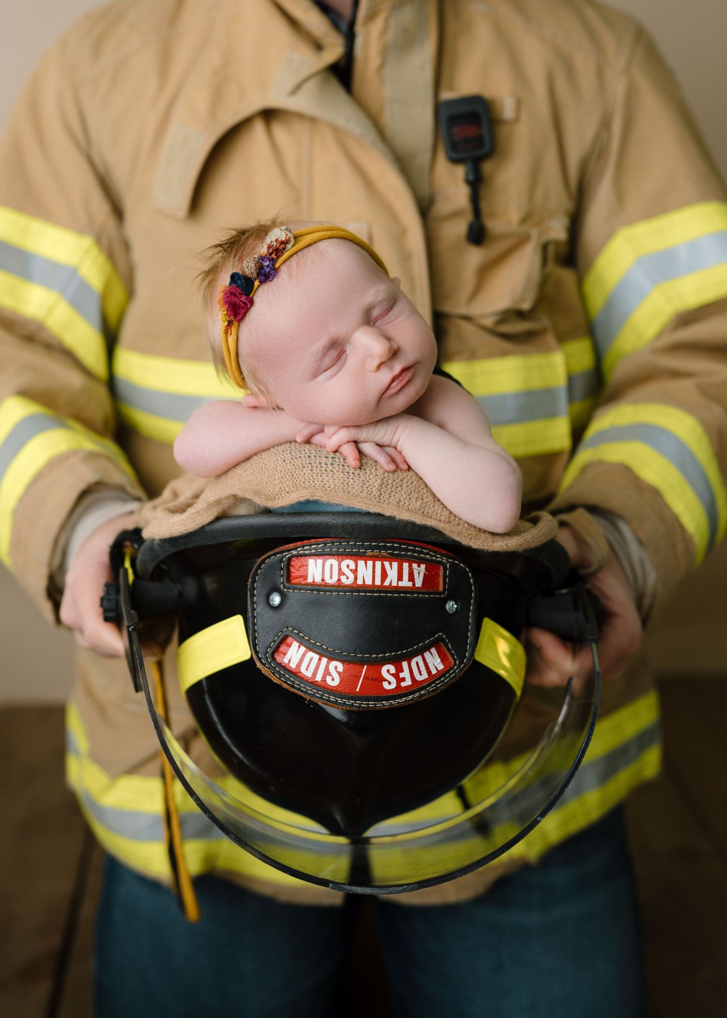 Sleeping newborn baby resting on firefighter helmet held by parent in yellow turnout gear.