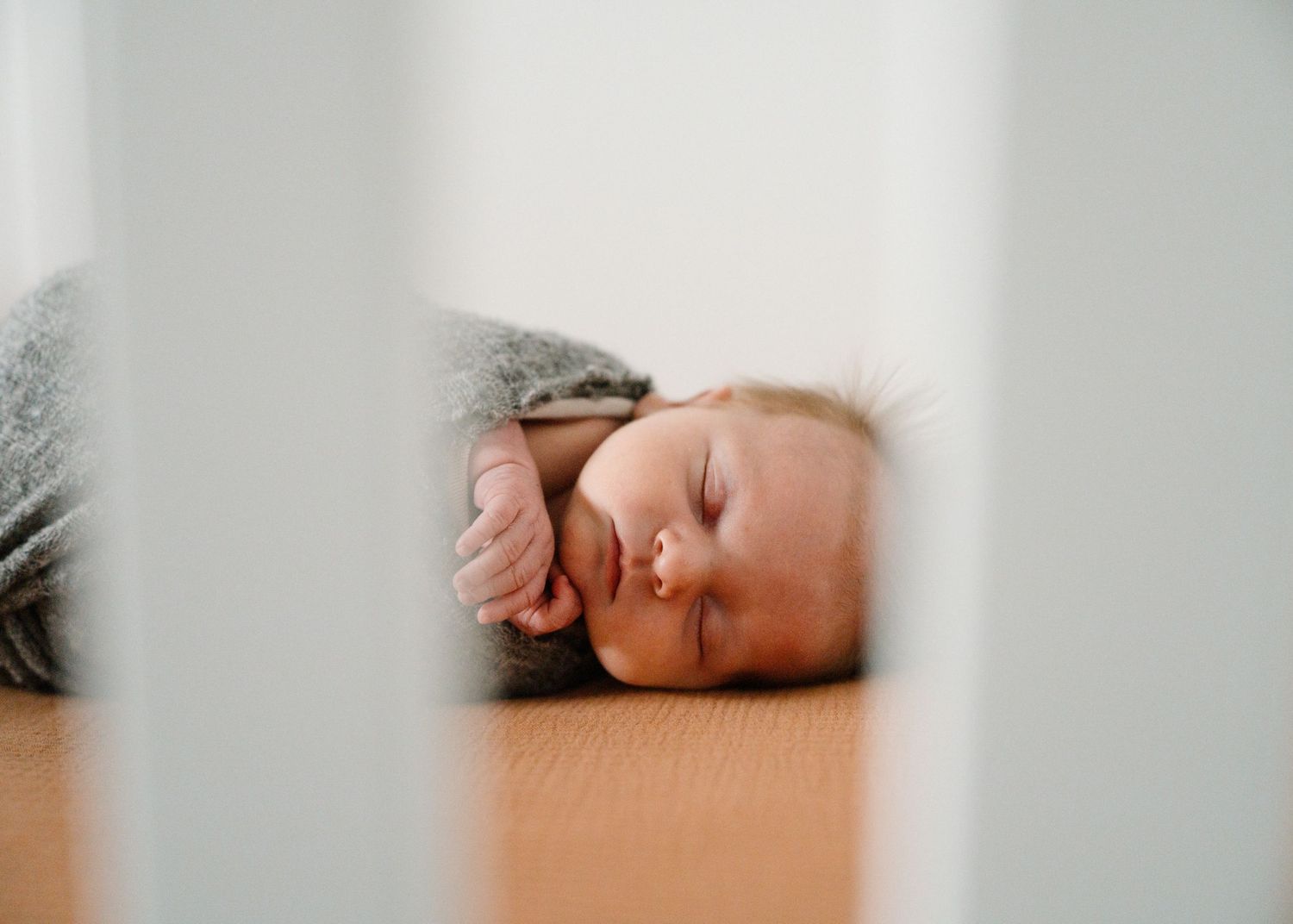 Peaceful newborn baby sleeping in gray swaddle viewed through white crib slats.
