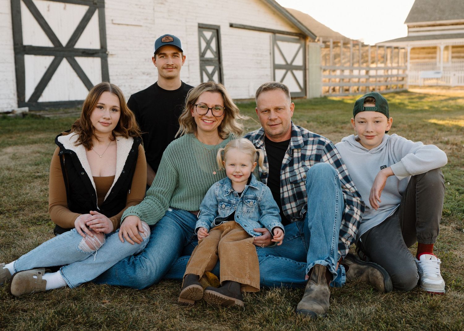 Extended family of six poses casually together on grass in front of a white barn at sunset.
