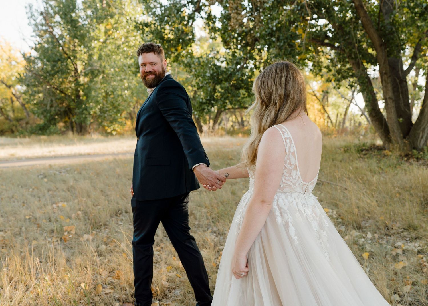 Groom in navy suit smiling as he leads bride in lace gown by the hand through a sunny meadow.