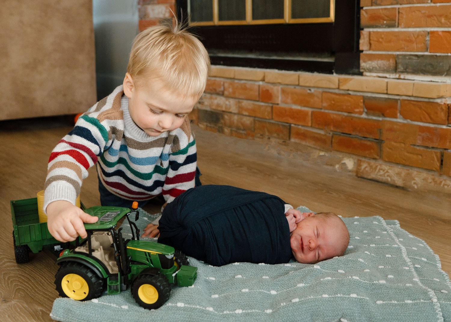 Young blonde toddler playing with green John Deere tractor beside sleeping swaddled newborn.