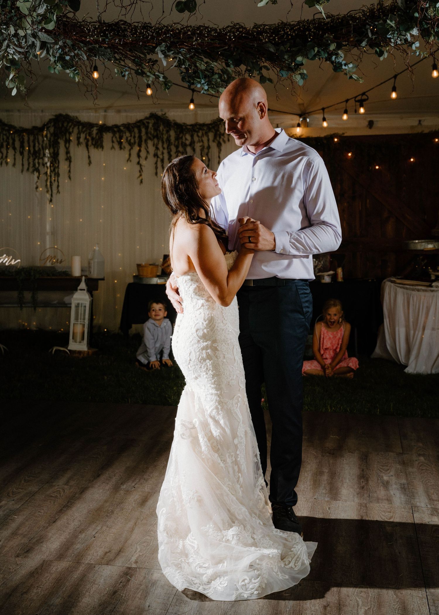 Bride and groom share their first dance under string lights and greenery at a romantic rustic wedding reception.