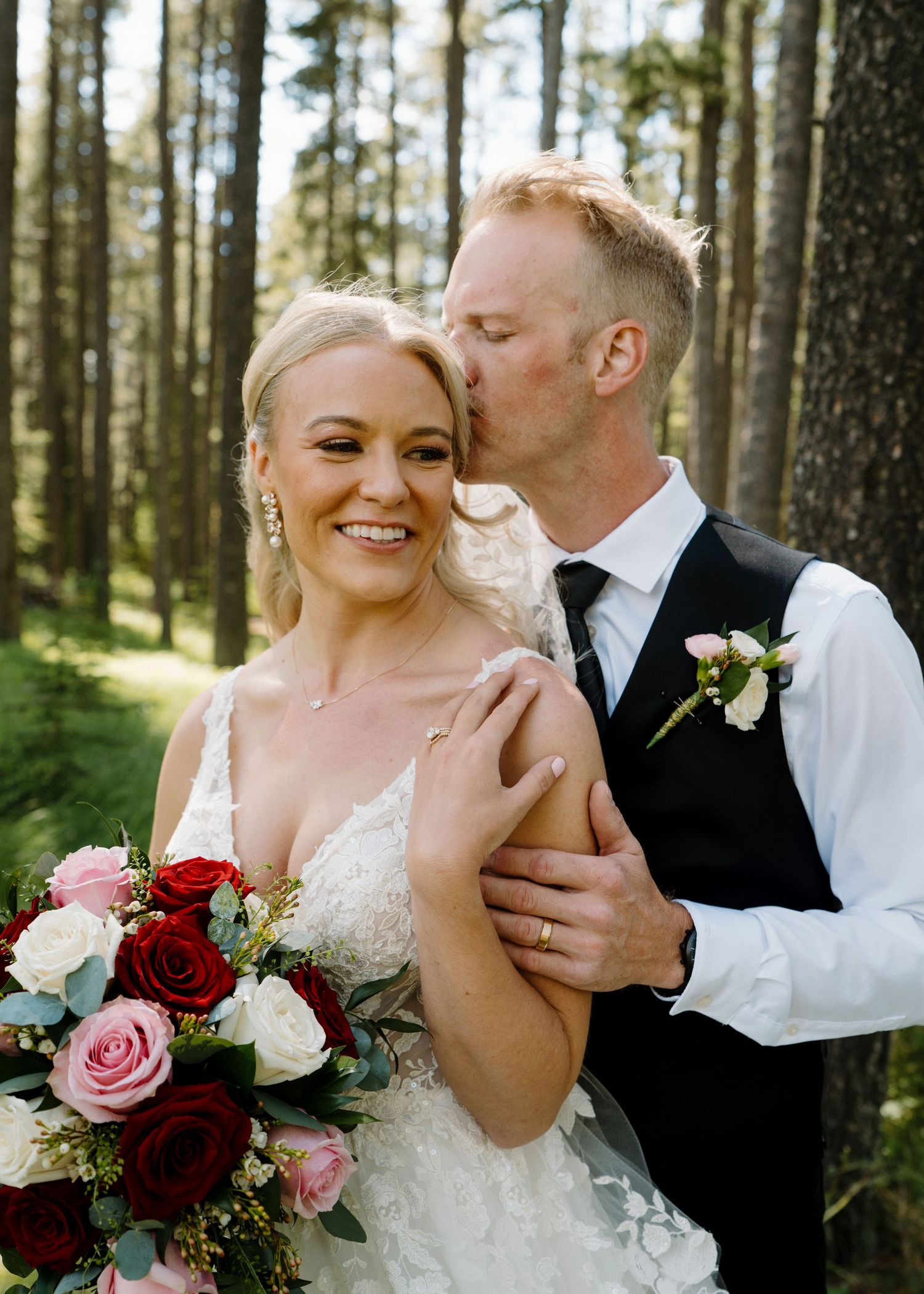 Bride holding red and pink rose bouquet smiles as groom kisses her cheek in a forest wedding portrait.