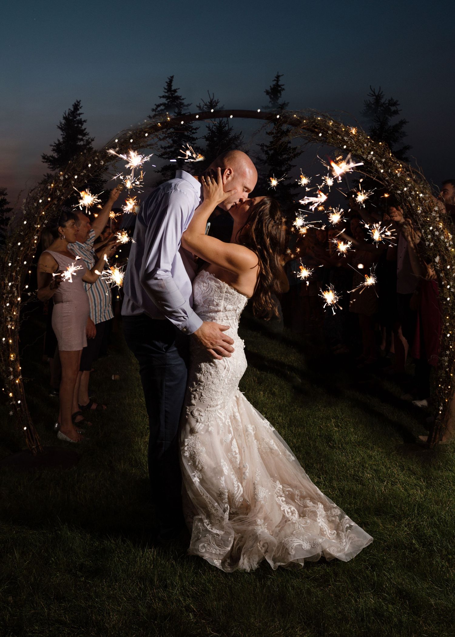Bride and groom share romantic kiss under glowing sparkler arch surrounded by wedding guests at night.