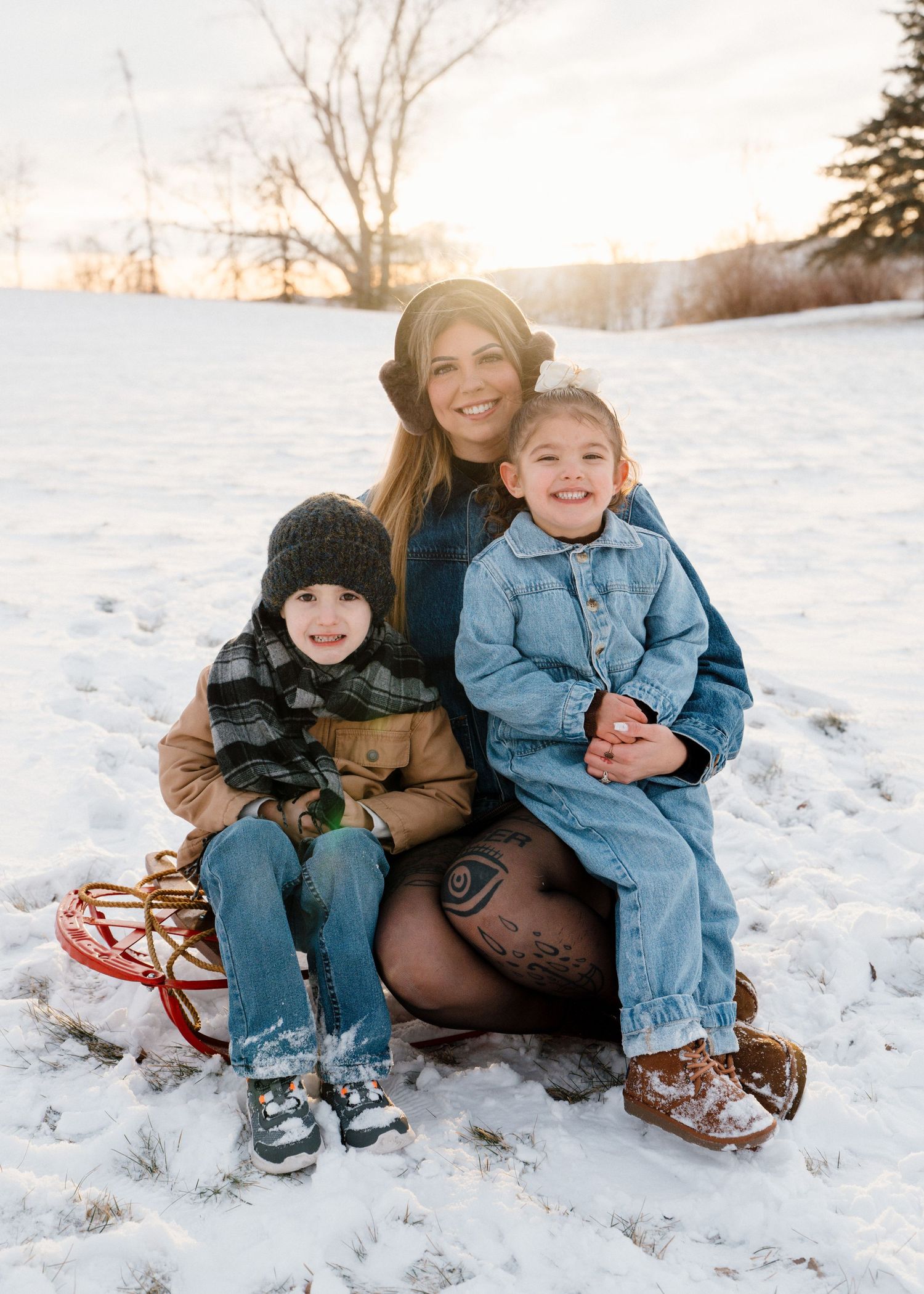 Mother sitting on sled in snow with two young children at golden hour sunset.